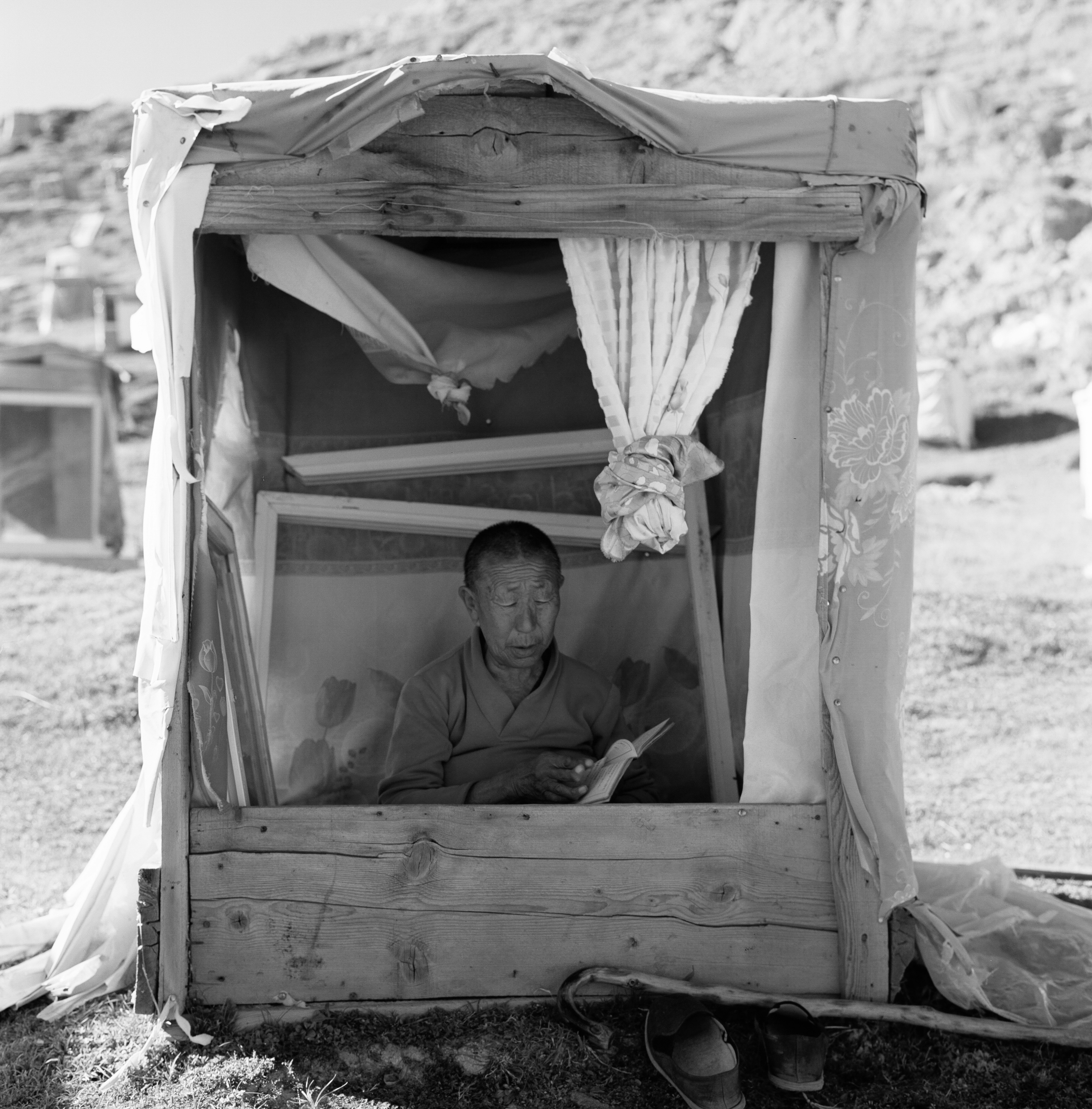 小屋の中に座っている男性のモノクロ写真 簡素な木製の構造とカーテンが特徴