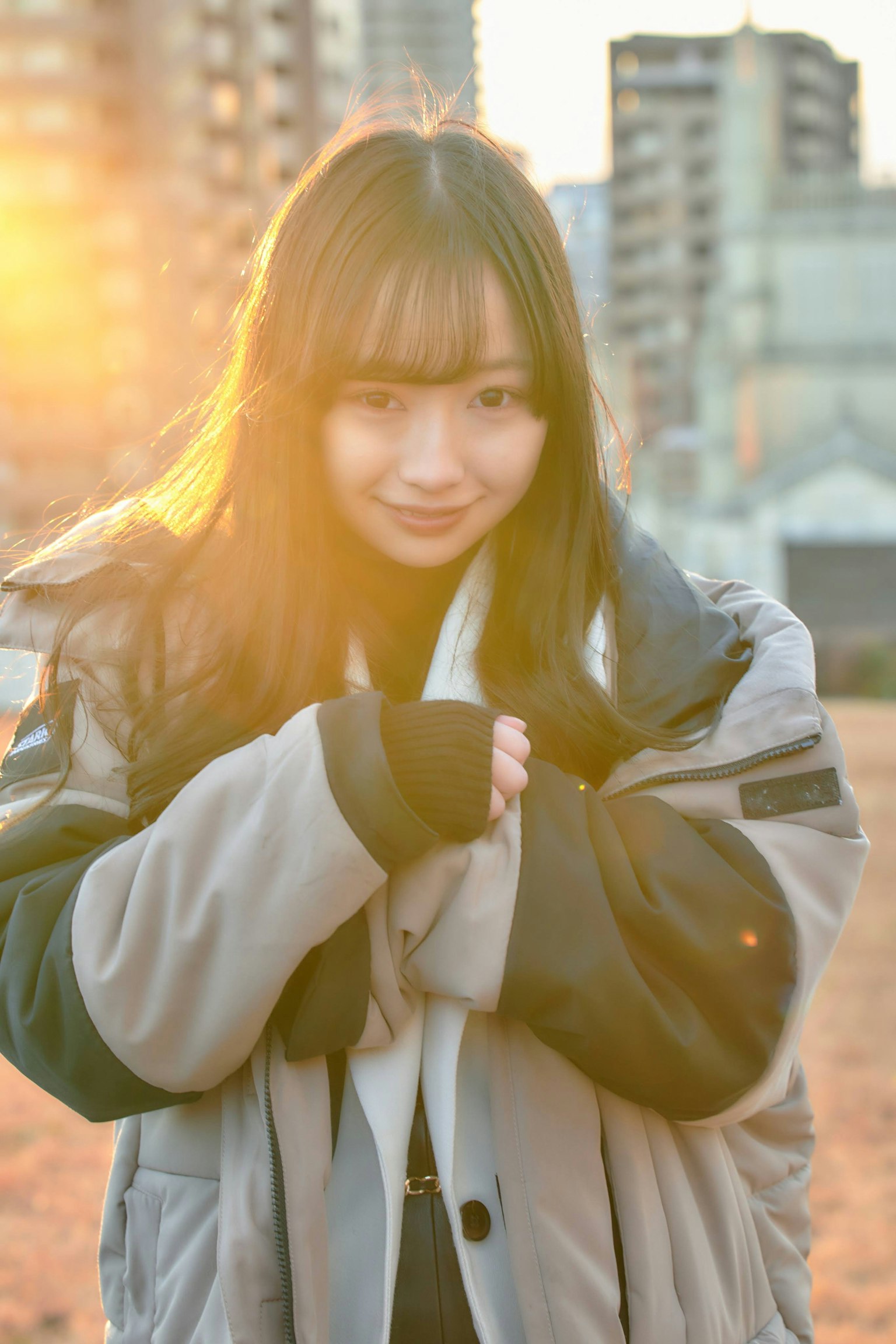 ภาพพอร์ตเทรตของผู้หญิงที่มีพระอาทิตย์ตกเป็นพื้นหลัง สวมเสื้อโค้ทหนา มือประสานกันและยิ้ม