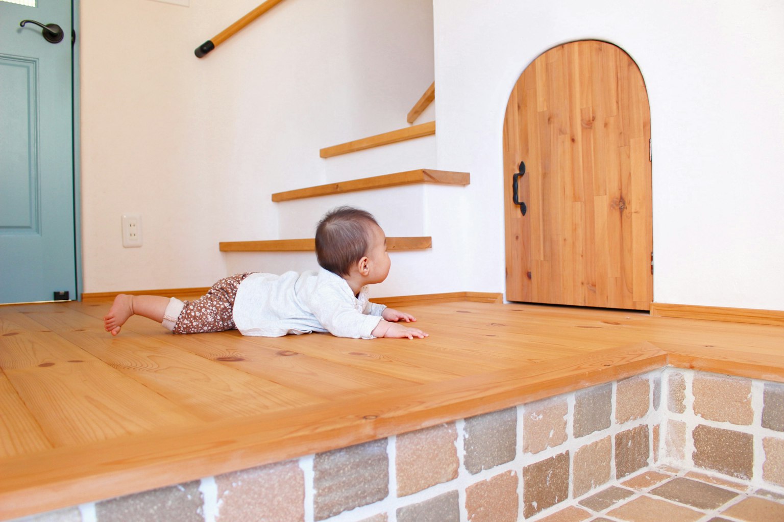 A baby crawling on a wooden floor towards a small door near stairs