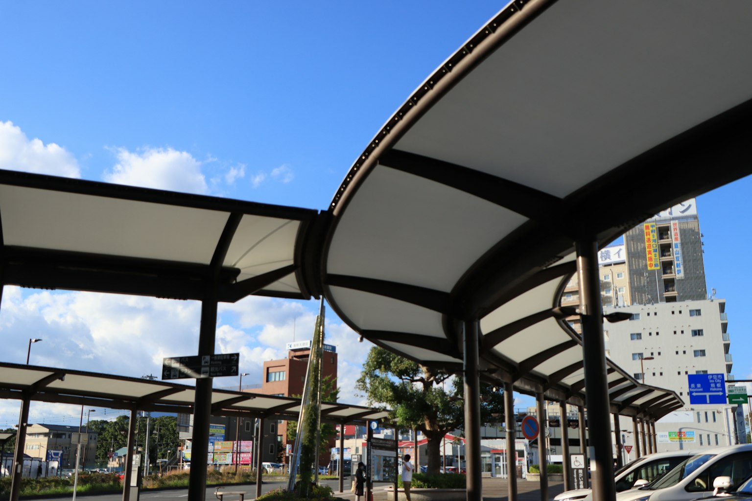 Modern bus stop canopies with curved roofs under a blue sky and surrounding buildings