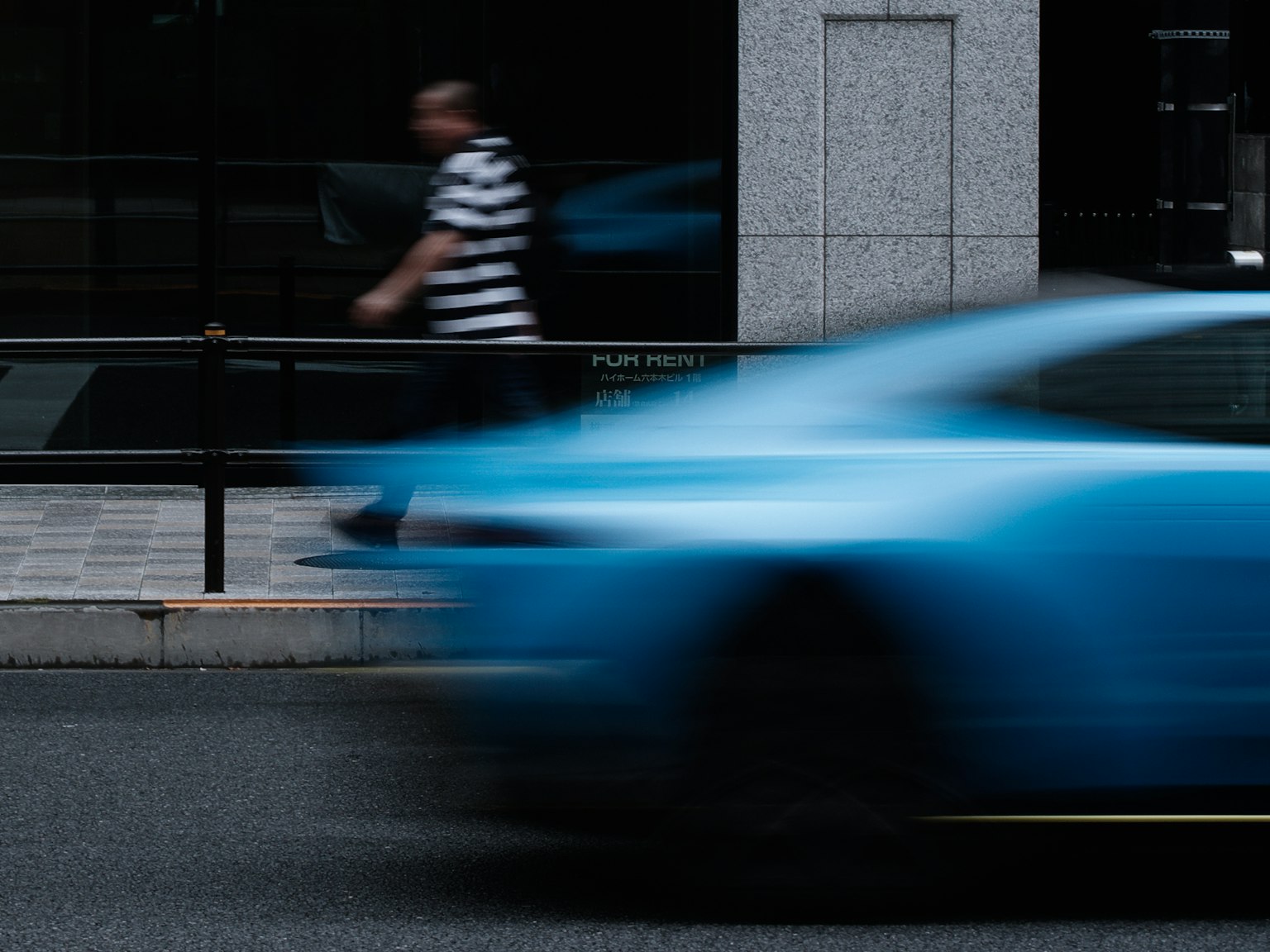 Un coche azul pasa rápidamente mientras una persona con una camisa a rayas camina por la calle