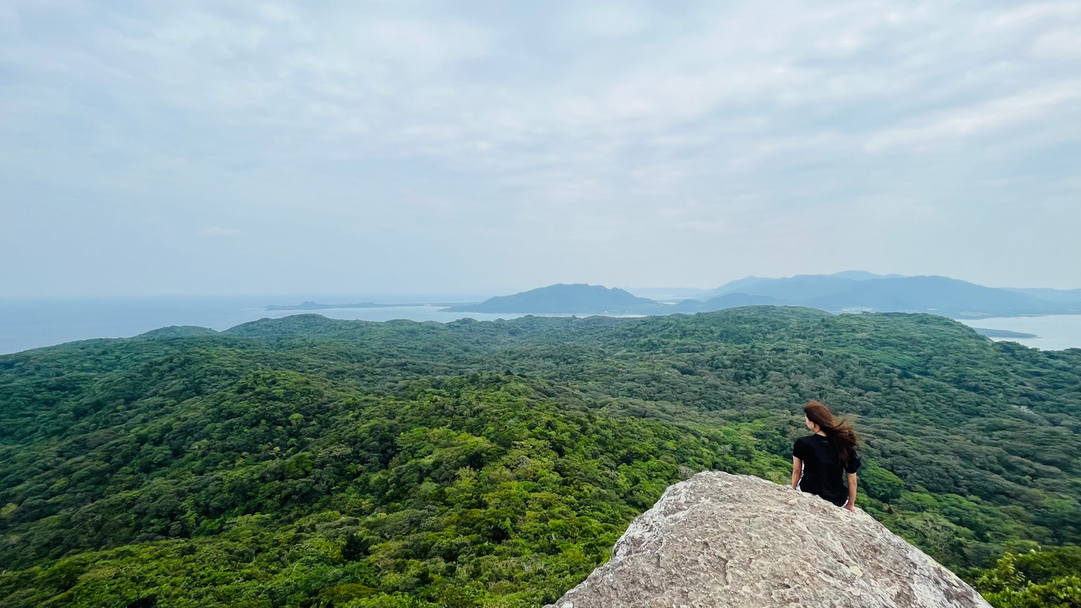 Woman sitting on a rocky ledge overlooking lush green hills and the sea