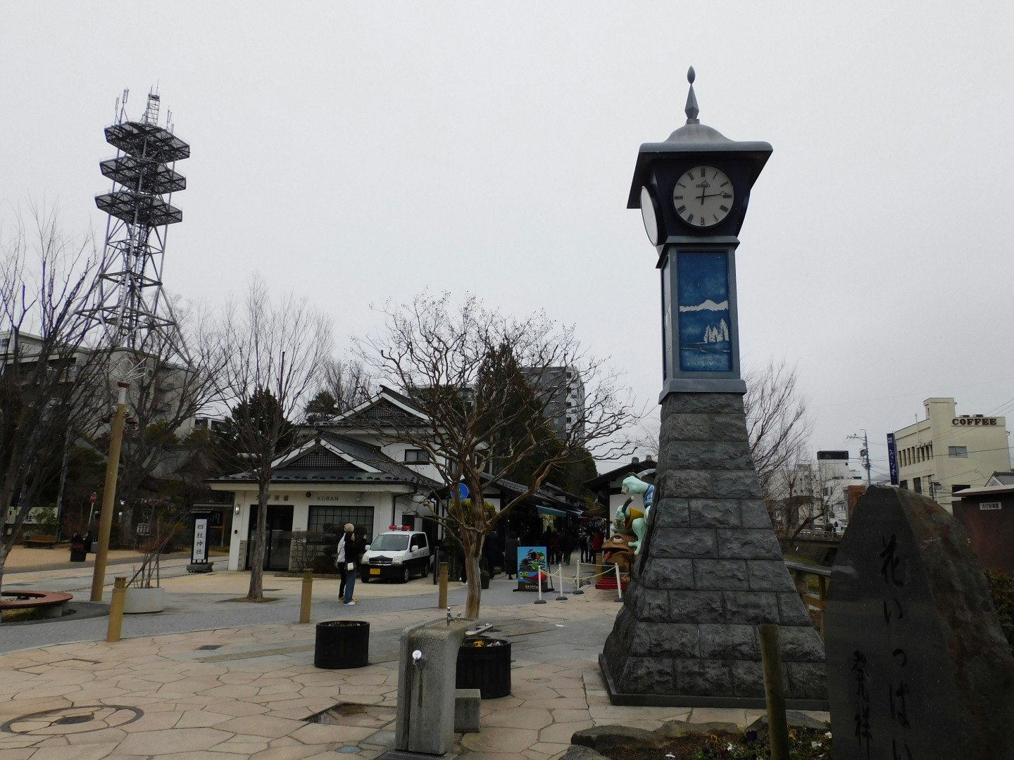 広場に立つ時計塔と送電鉄塔のある街並み