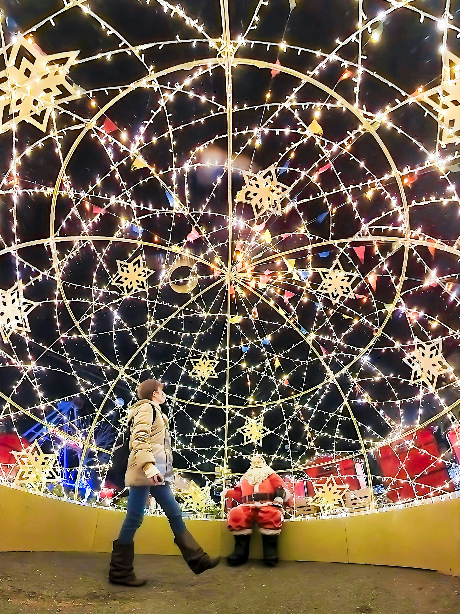 Một người phụ nữ đi gần một đứa trẻ ngồi bên cạnh ông già Noel được bao quanh bởi những ánh đèn Giáng sinh lấp lánh