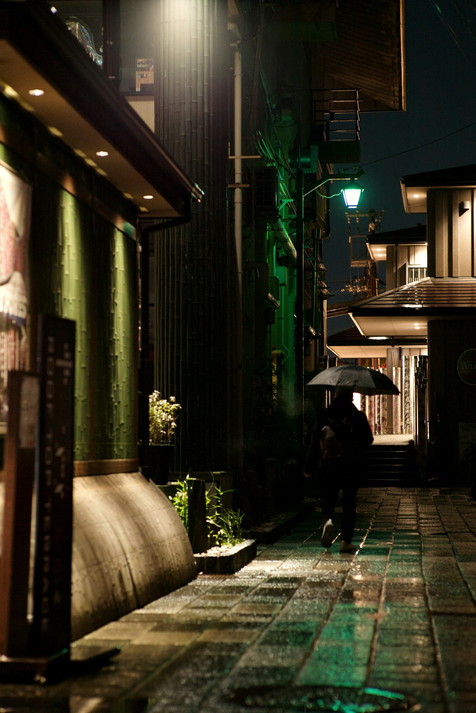 A quiet night alley featuring a person with an umbrella and distinctive green lights