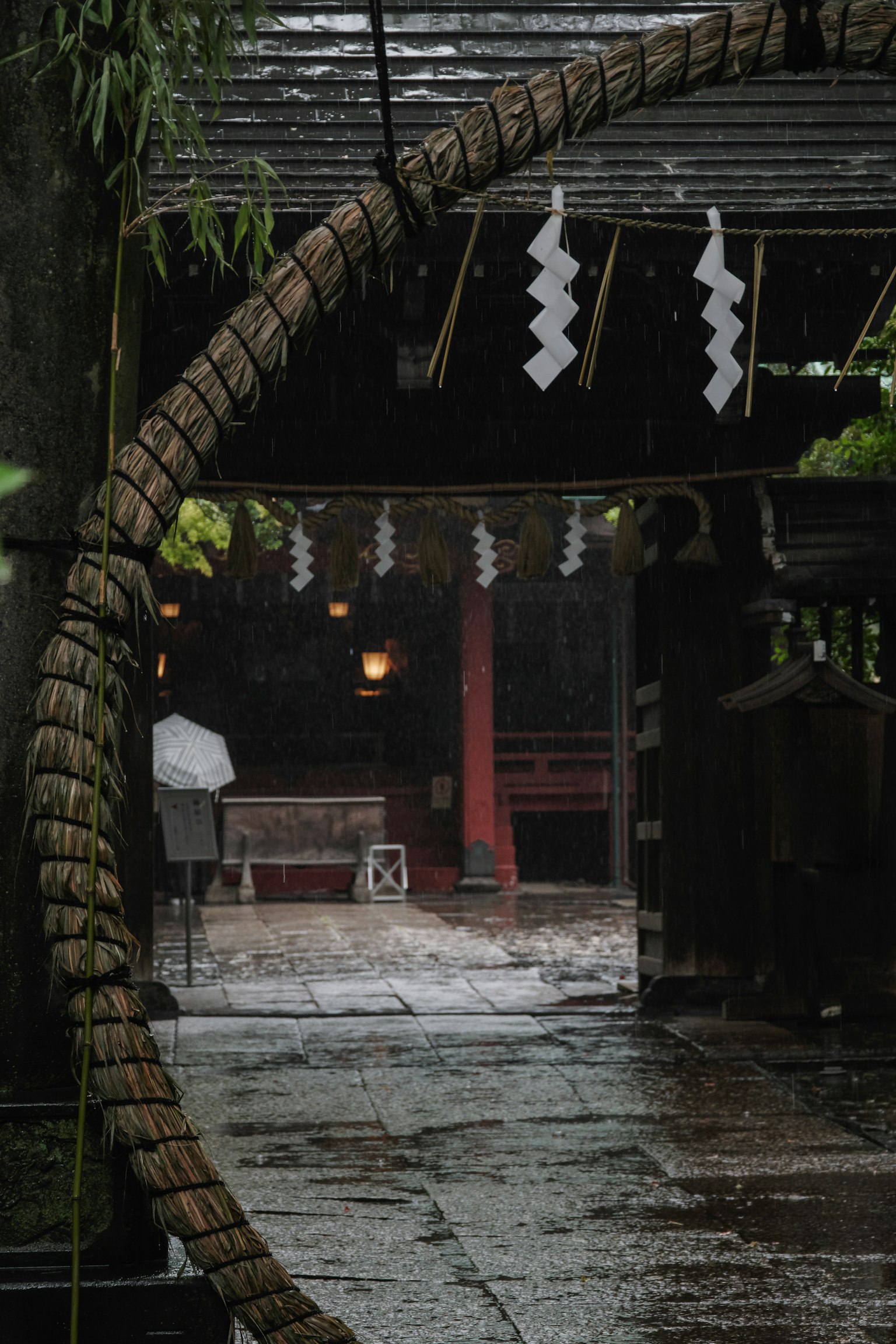 Entrance of a shrine adorned with a large straw ring and white decorations in the rain