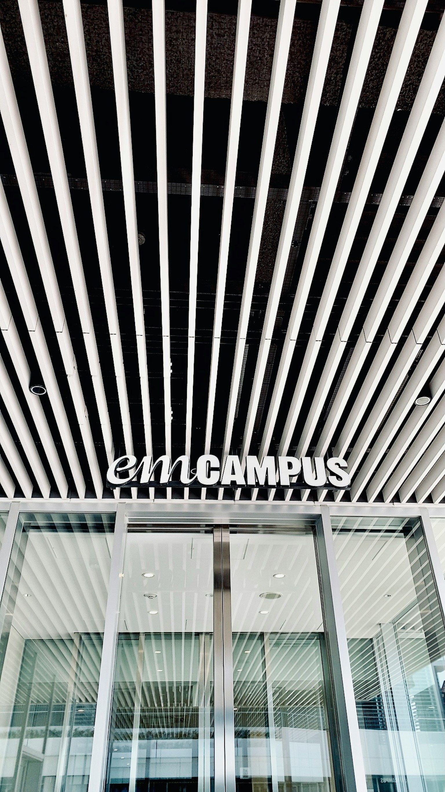 Bên ngoài tòa nhà hiện đại với trần sọc trắng lối vào kính logo emCAMPUS