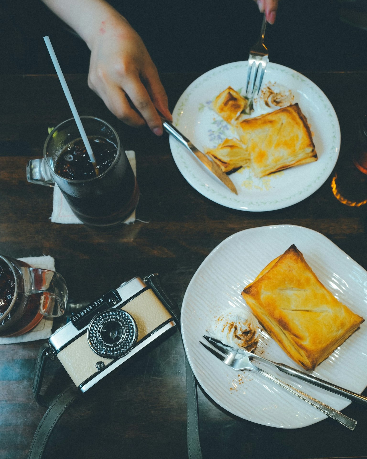 Una mano sosteniendo utensilios sobre un plato con postre y bebidas en una mesa