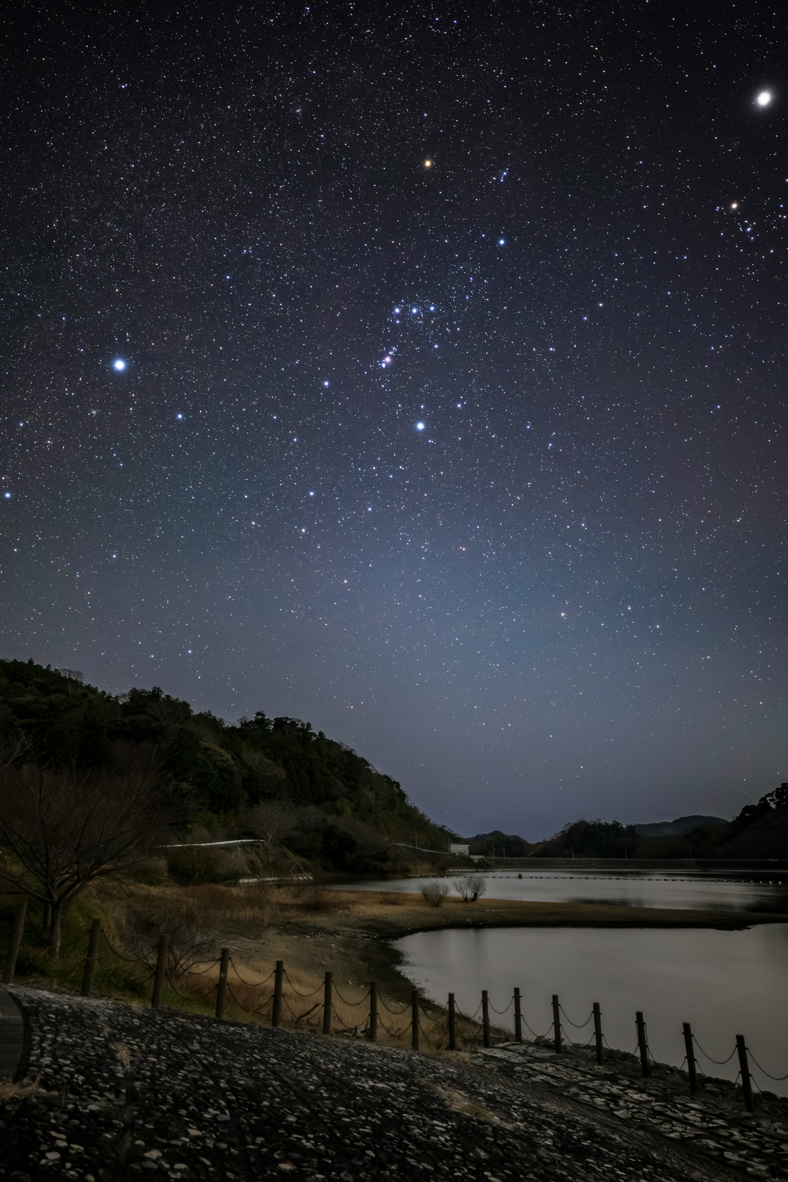 Starry night sky over a calm water surface with silhouettes of hills