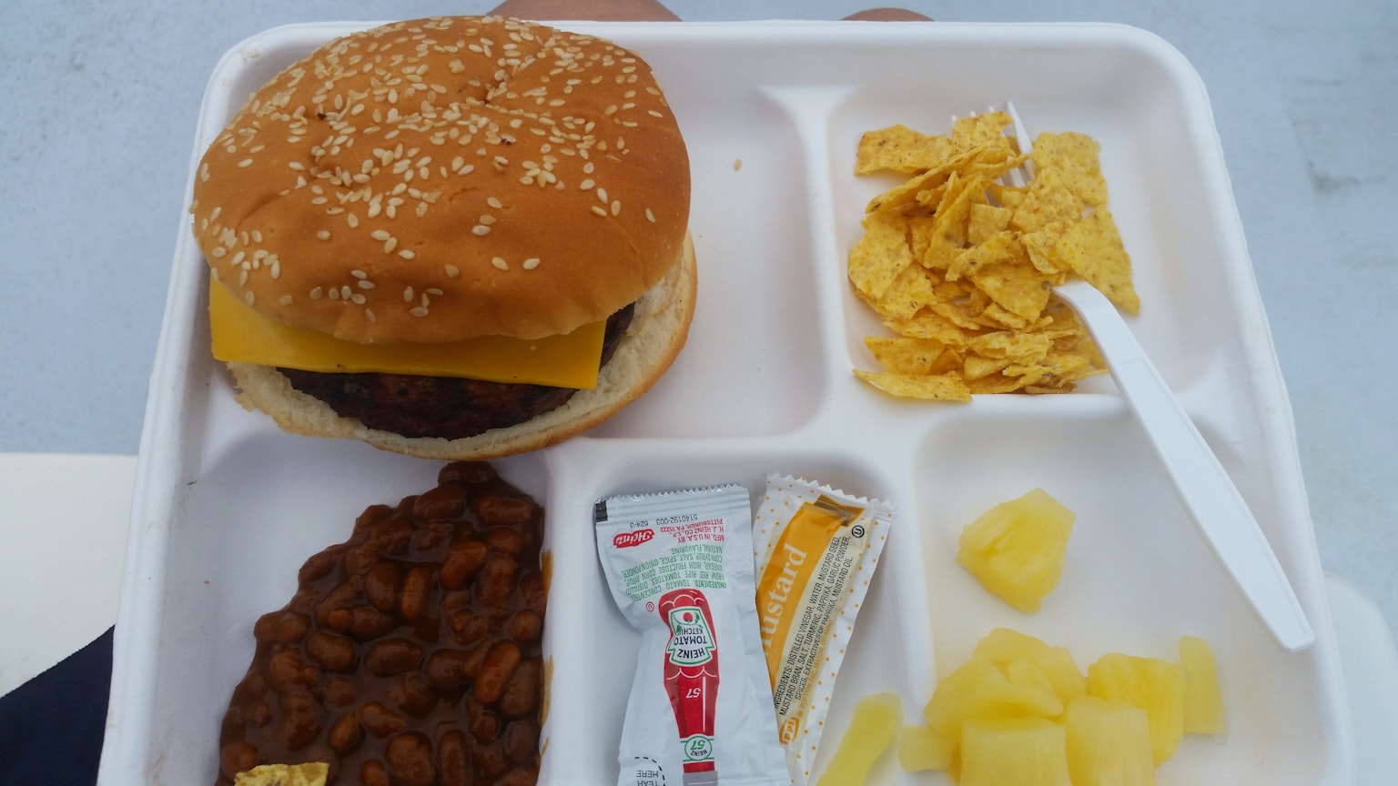 Cheeseburger mit Sesambrötchen, dazu Bohnen, Ananas, Cornflakes und Sauce auf einem Tablett