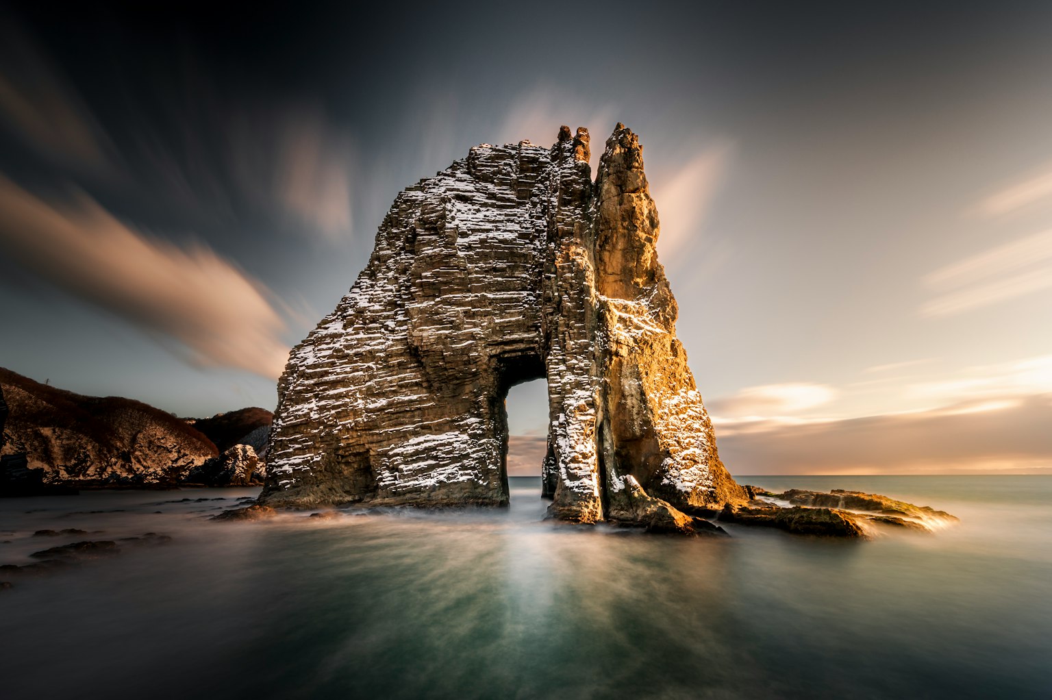 Ein großer Felsenbogen, der aus dem Meer ragt, mit einem dramatischen Sonnenuntergangshimmel