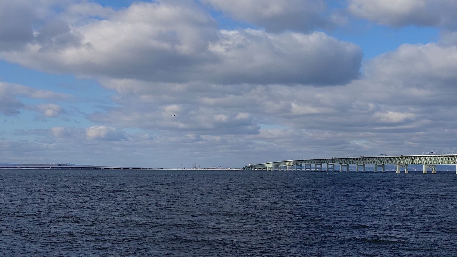 A scenic view of blue water with white clouds and a bridge in the distance