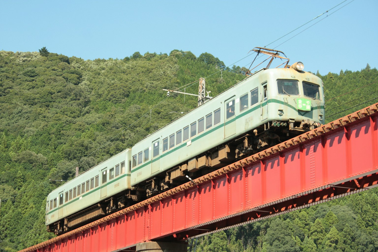 Ein grüner Zug überquert eine rote Brücke mit grünen Bergen im Hintergrund
