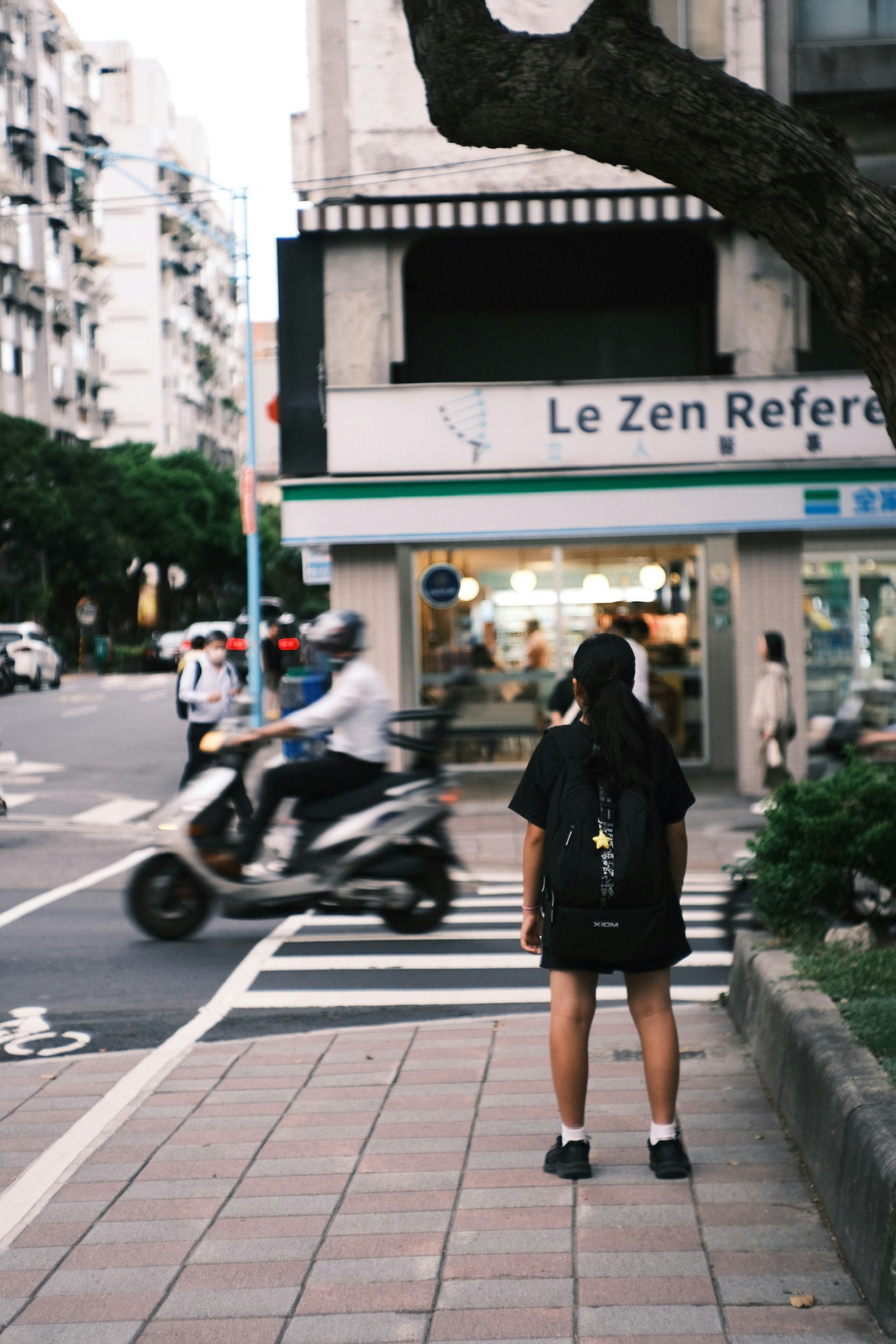 城市環境中一個人站在斑馬線上 一輛摩托車經過 一個寫著'Le Zen Refered'的商店招牌