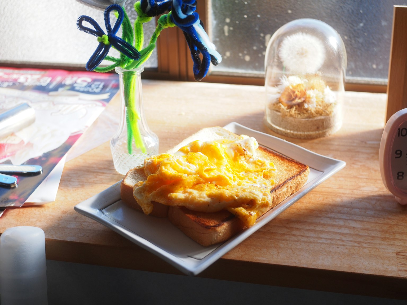 窓辺に置かれたトーストと卵料理の皿 花瓶と時計が背景にある