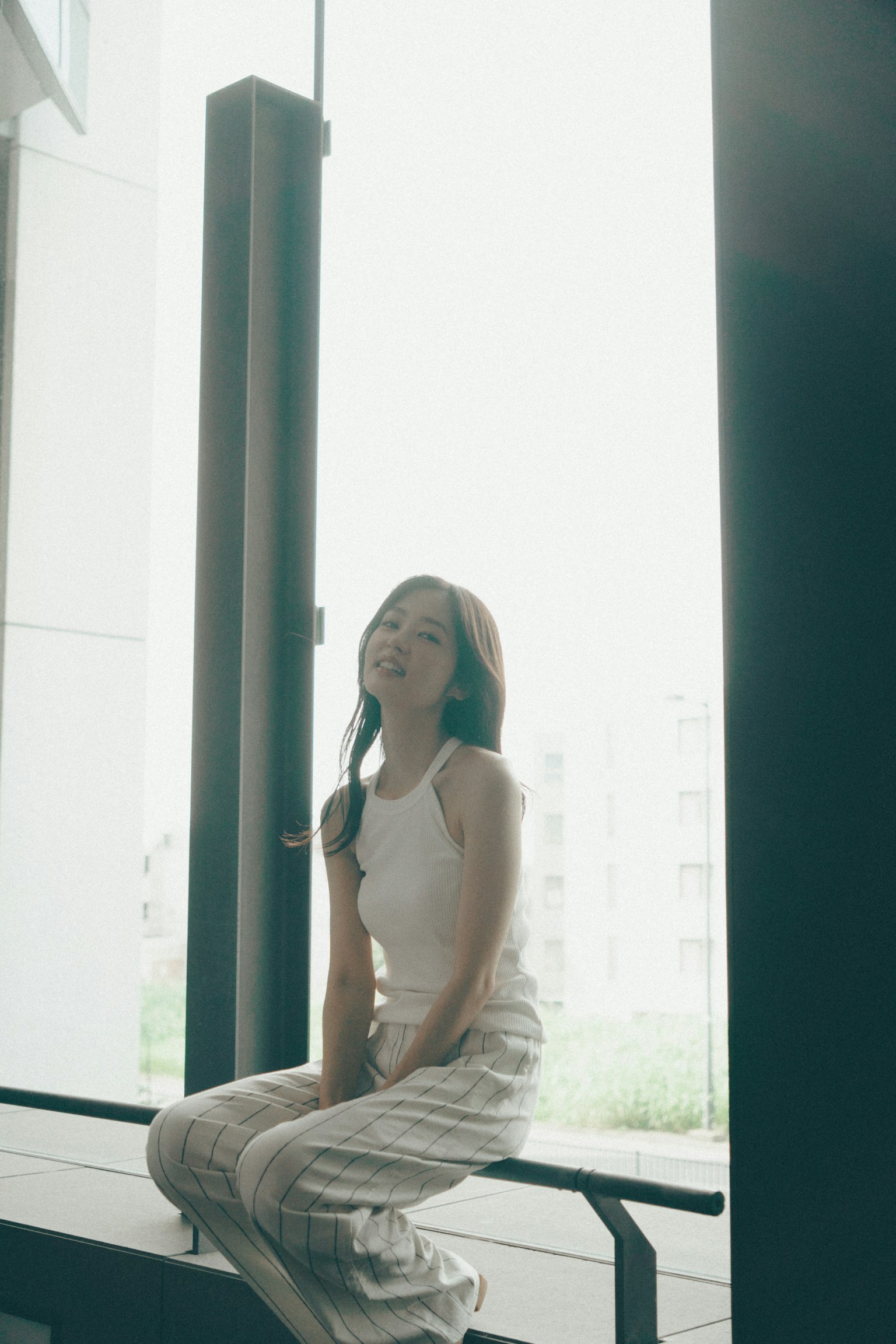 Mujer sentada junto a la ventana con una blusa y pantalones blancos, luz natural que entra