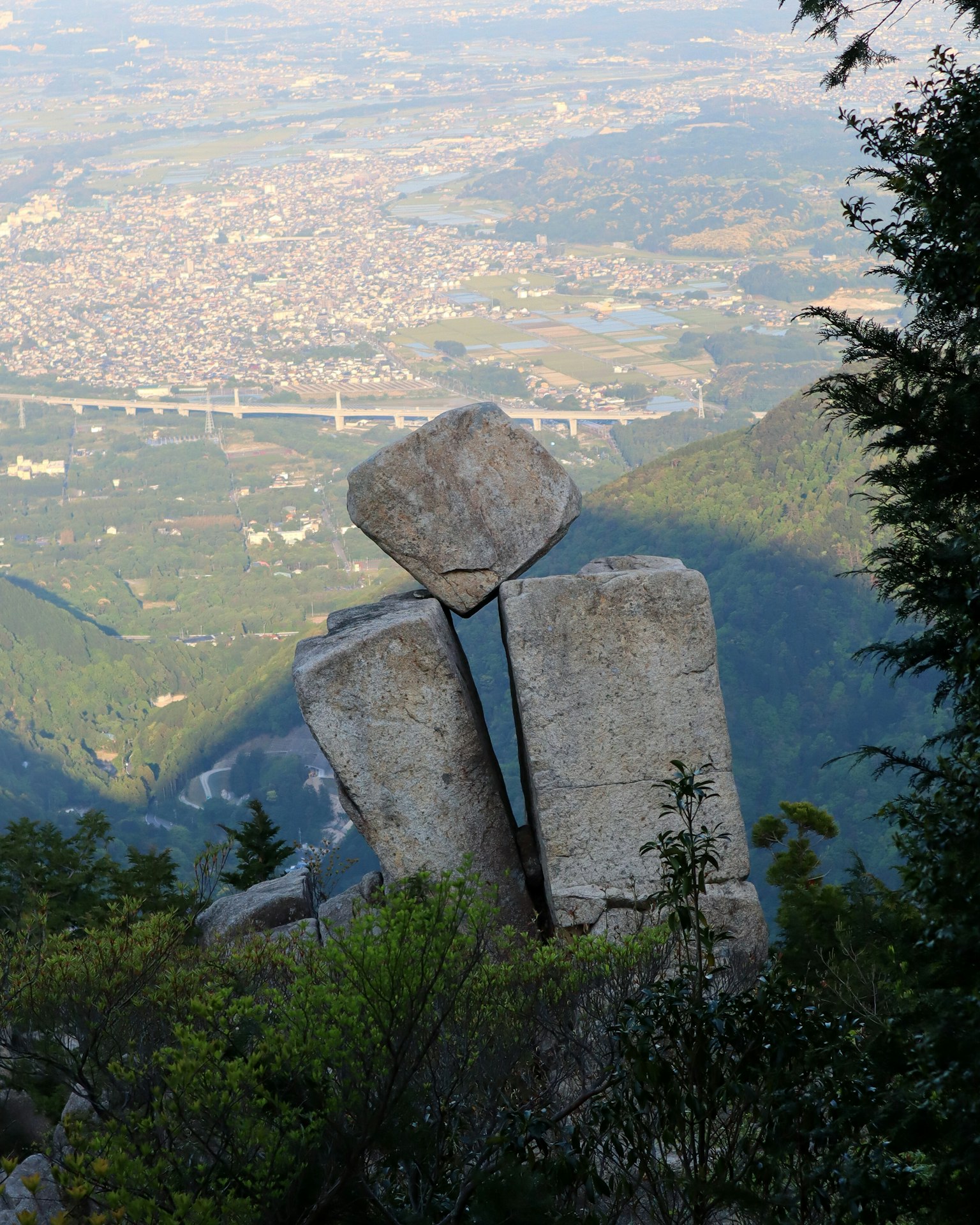 Two large stones forming a sculpture at the mountain top with a small stone between them surrounded by greenery and a scenic mountain view