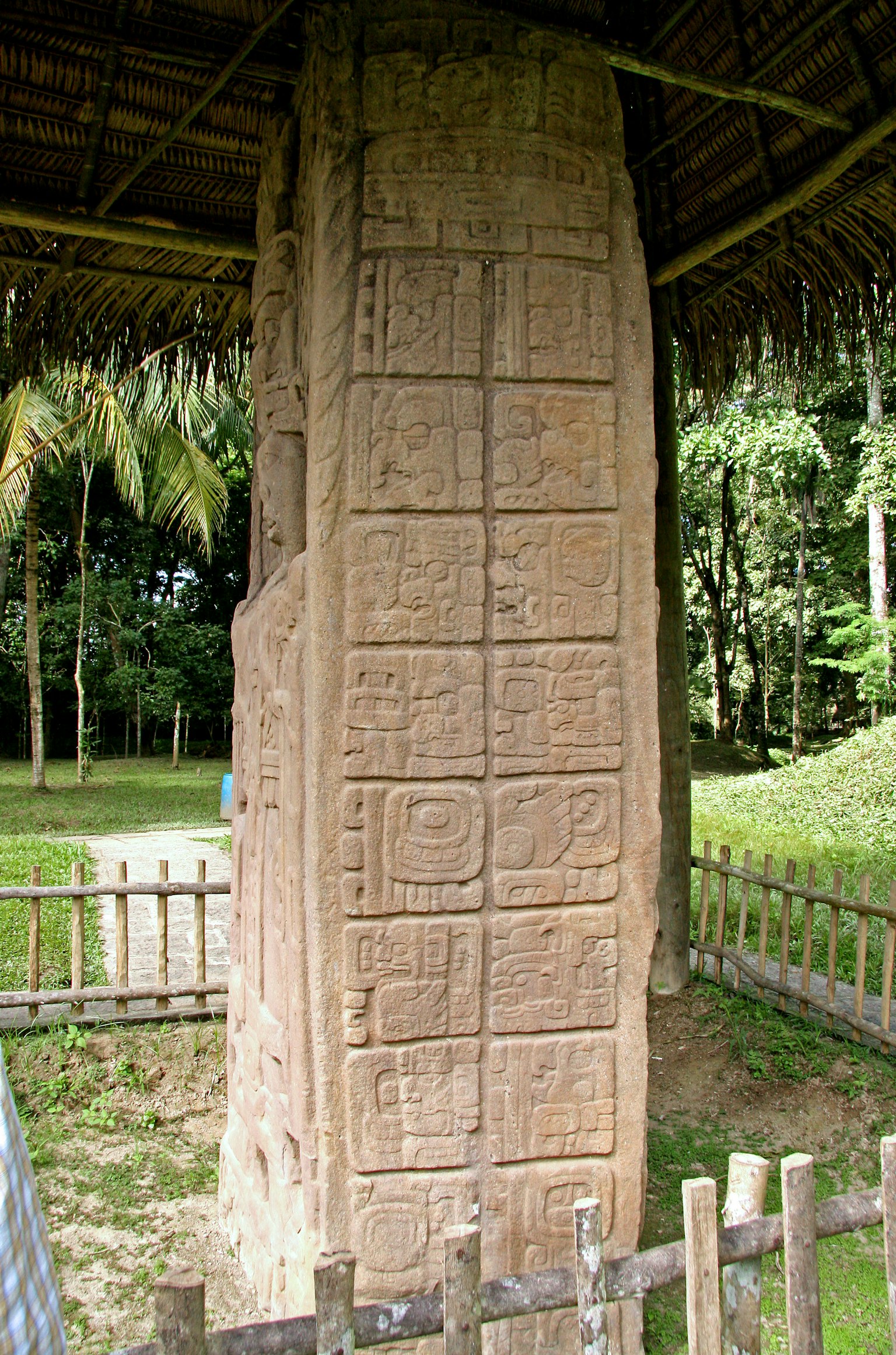 Monumen batu kuno dengan ukiran di bawah atap jerami dikelilingi oleh pepohonan hijau