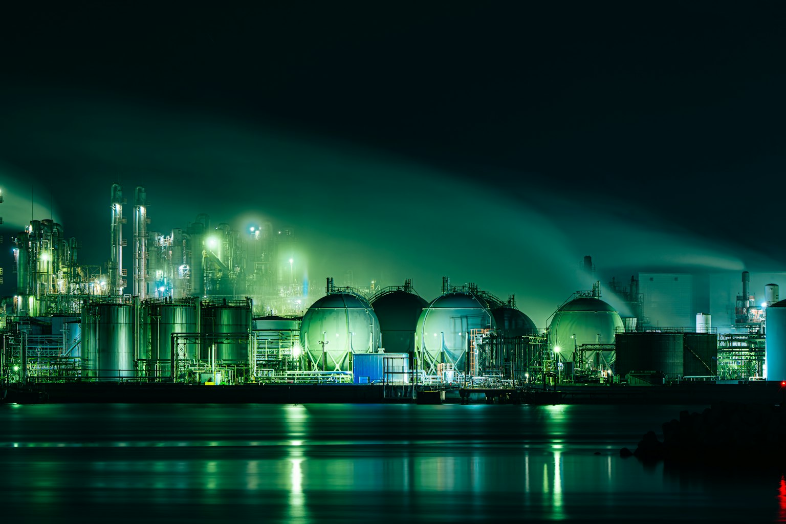 Vista nocturna de una instalación industrial iluminada en verde con grandes tanques esféricos