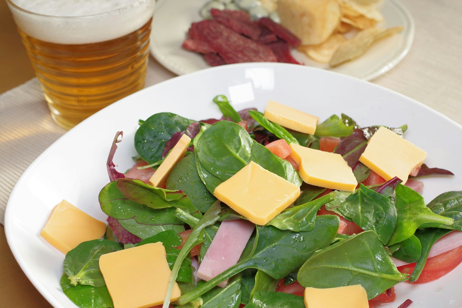 Một bát salad với rau xanh và phô mai, kèm theo một ly bia và một đĩa thịt và phô mai ở phía sau