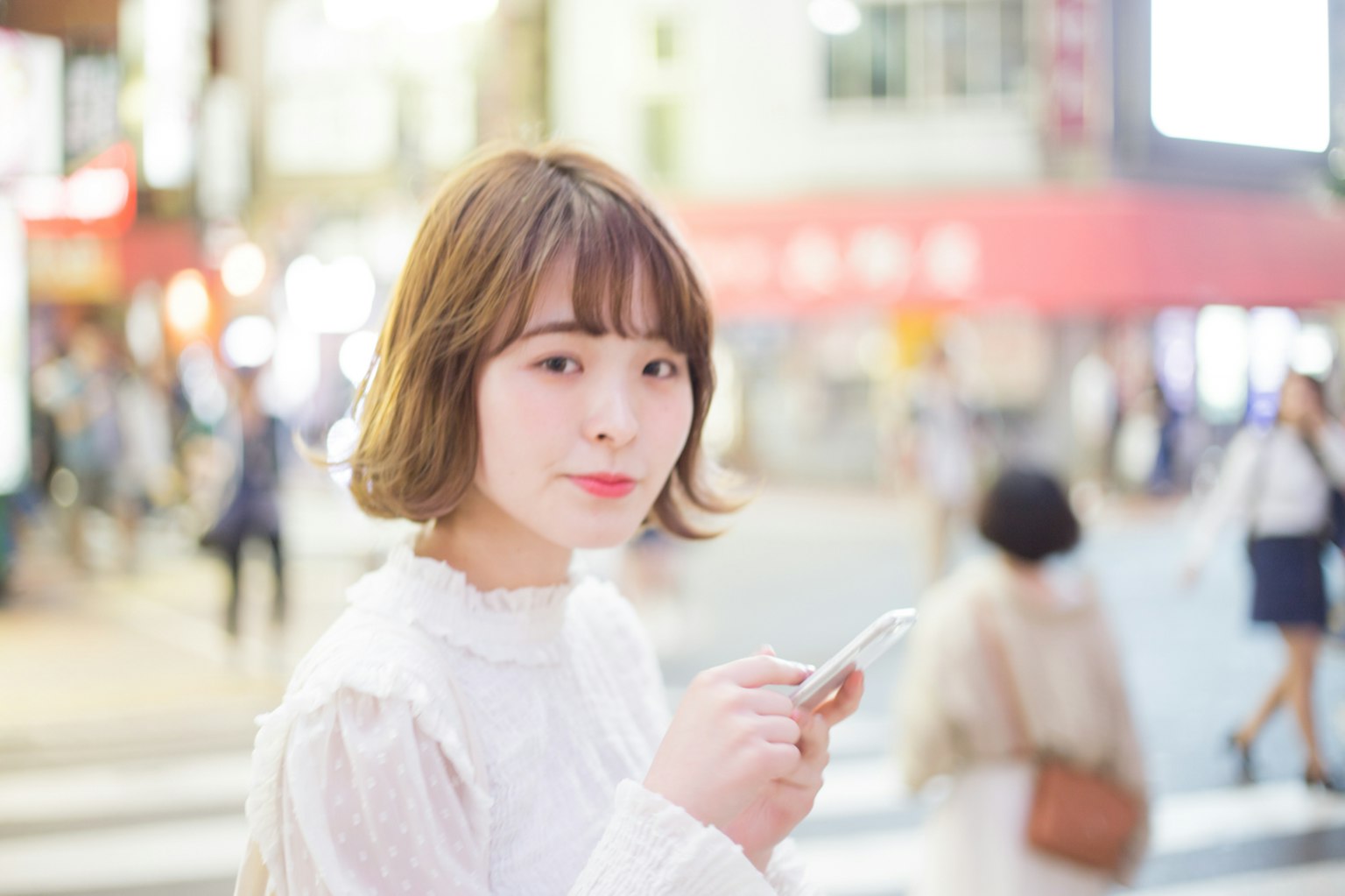 Portrait of a woman holding a smartphone at a street corner She is wearing a white outfit with blurred people in the background