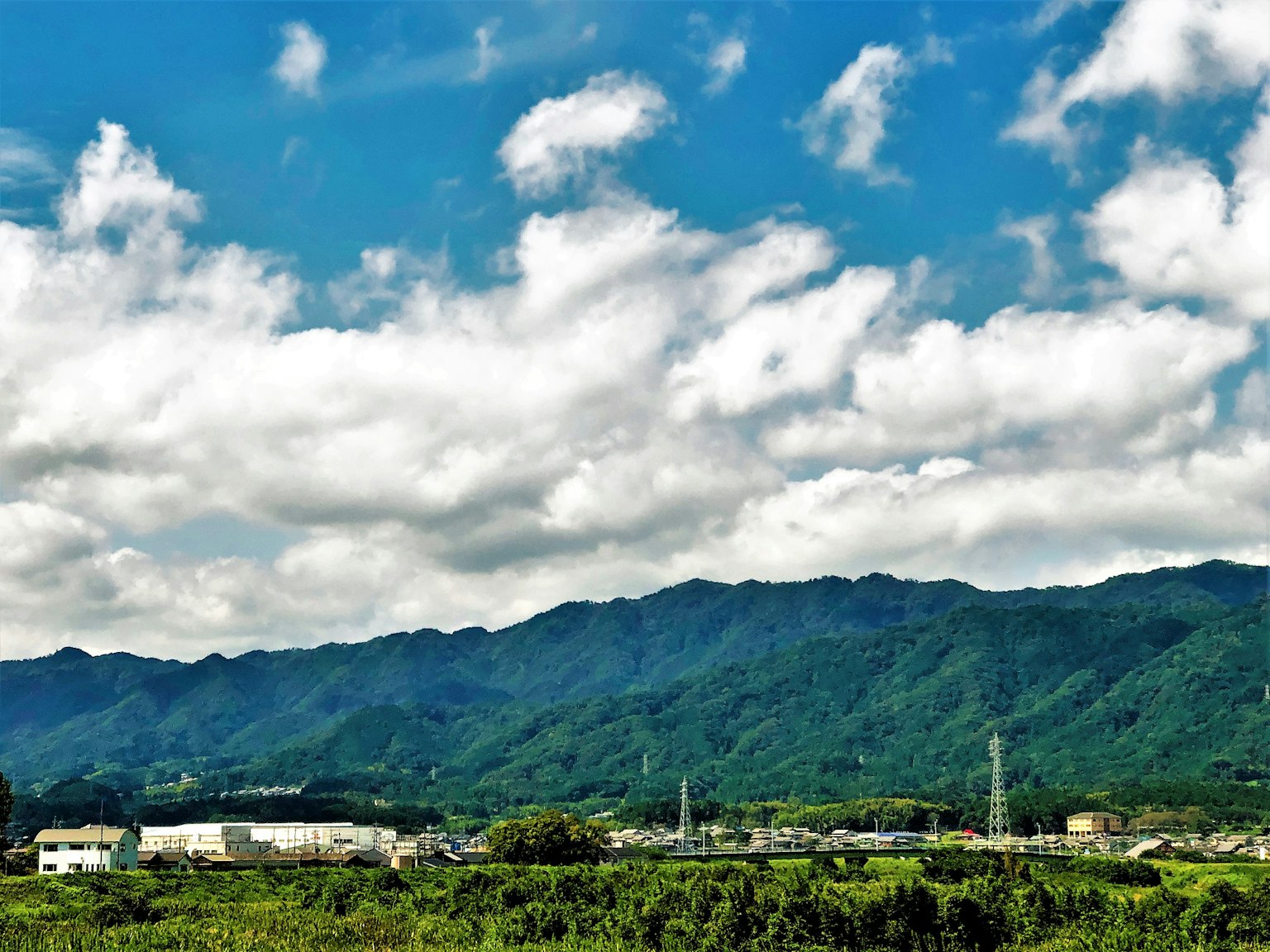 Landscape featuring green mountains under a blue sky with clouds and industrial buildings