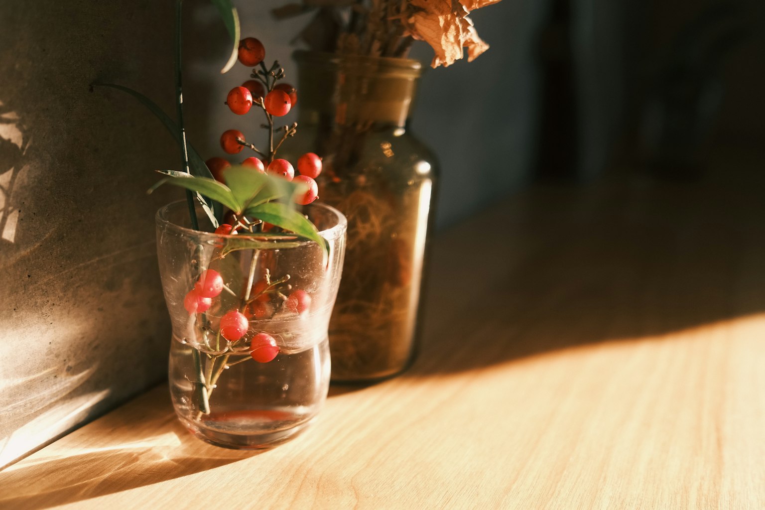 Un vaso con bayas rojas y un jarrón de flores secas sobre una mesa de madera