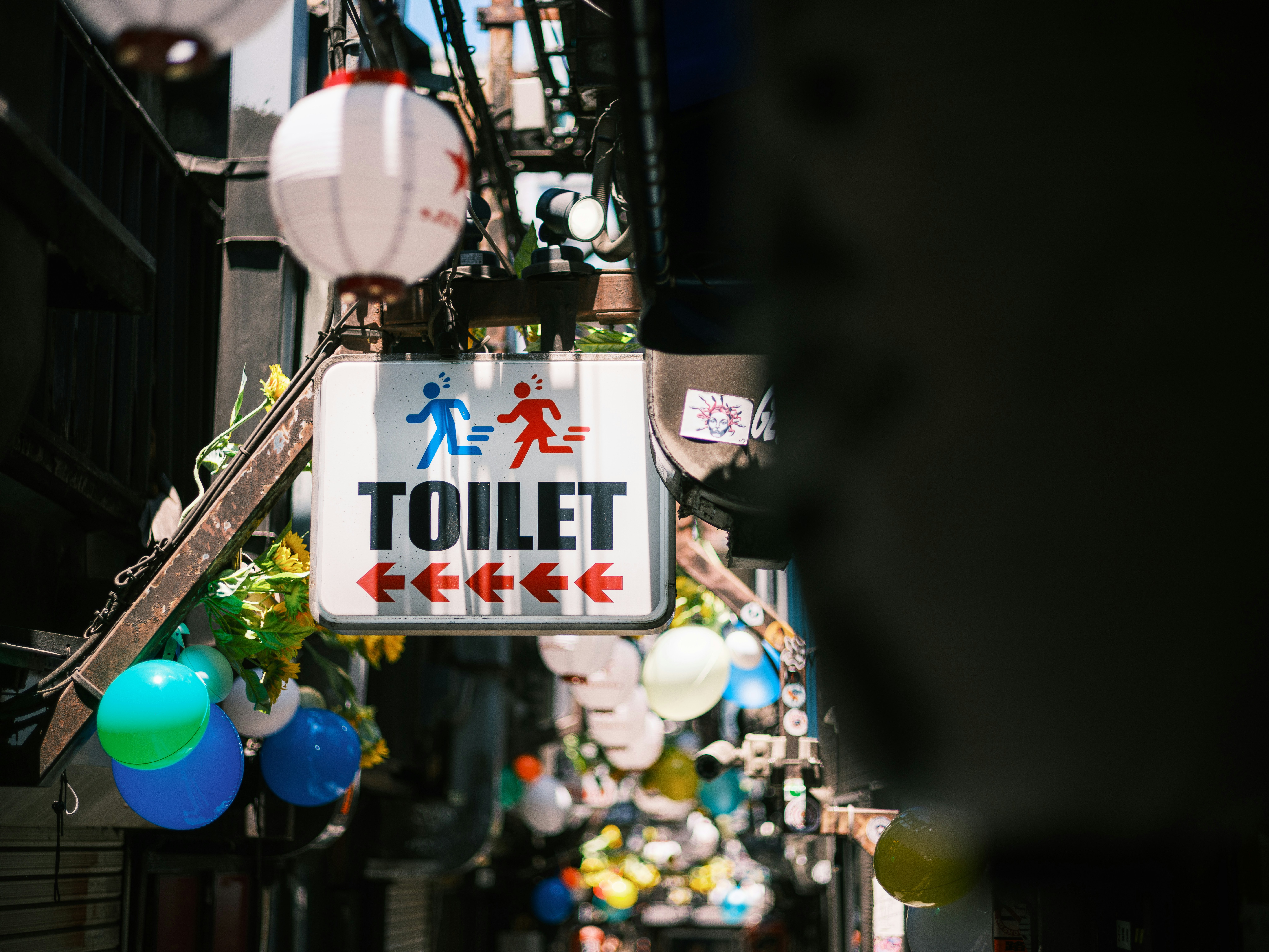 路地の天井に吊るされたカラフルな風船と「TOILET」標識のある案内板