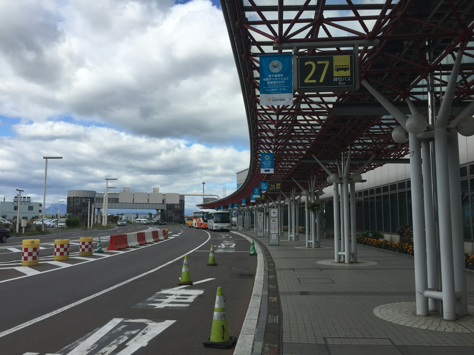 Tampilan luar area keberangkatan bandara dengan langit biru dan awan menunjukkan tanda 27 dan kerucut lalu lintas yang ditempatkan di sepanjang jalan