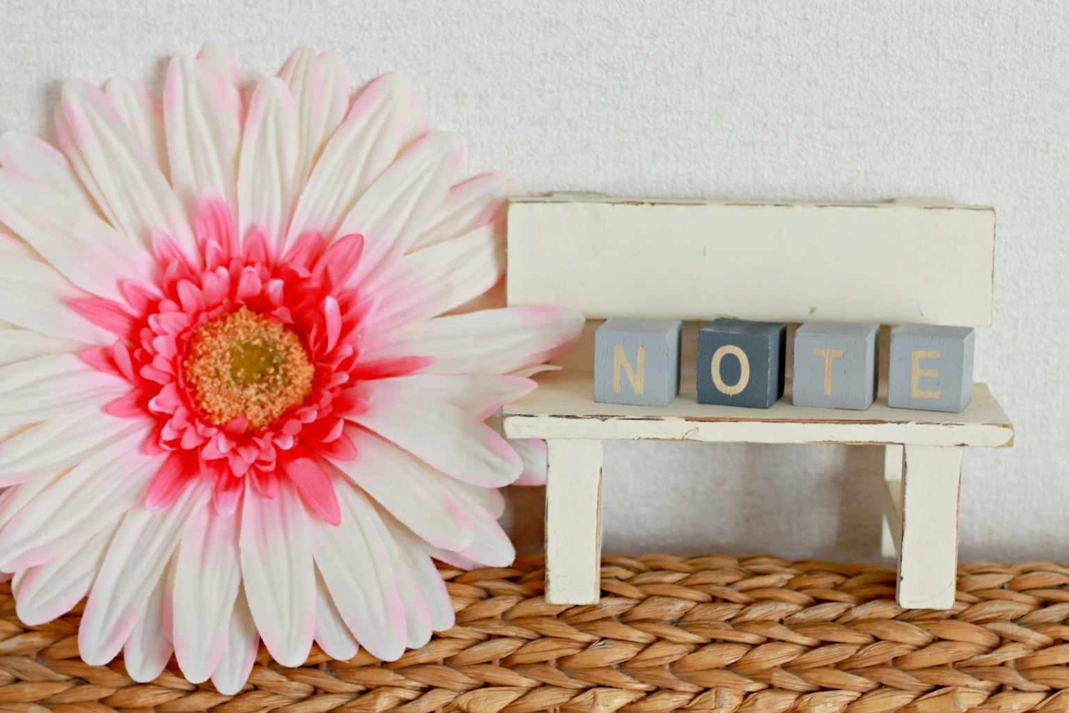 Un pequeño banco con bloques de madera que deletrean NOTE junto a una gran flor rosa contra una pared blanca