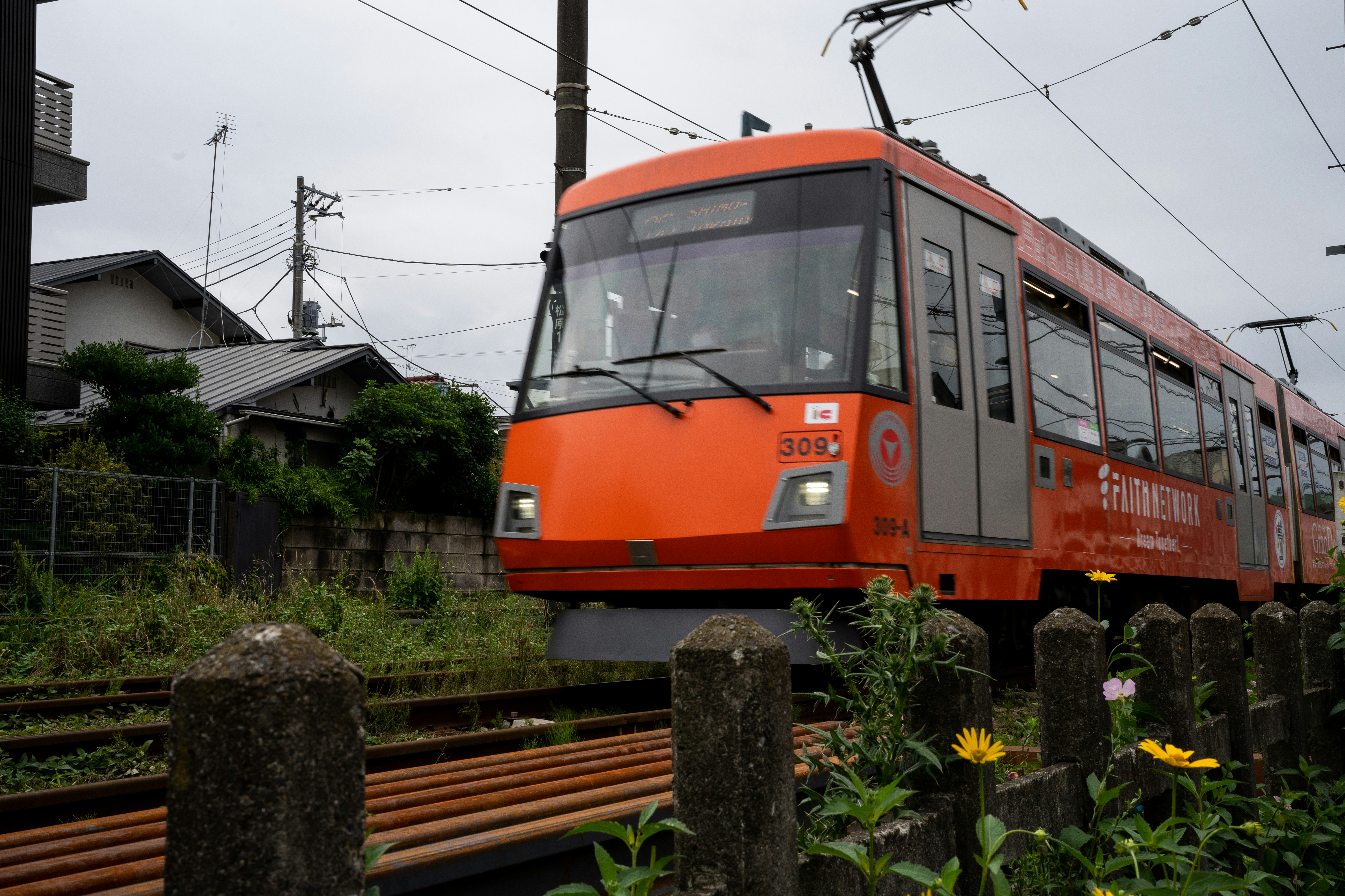 一列橙色火車在軌道上行駛，背景有房屋和植物