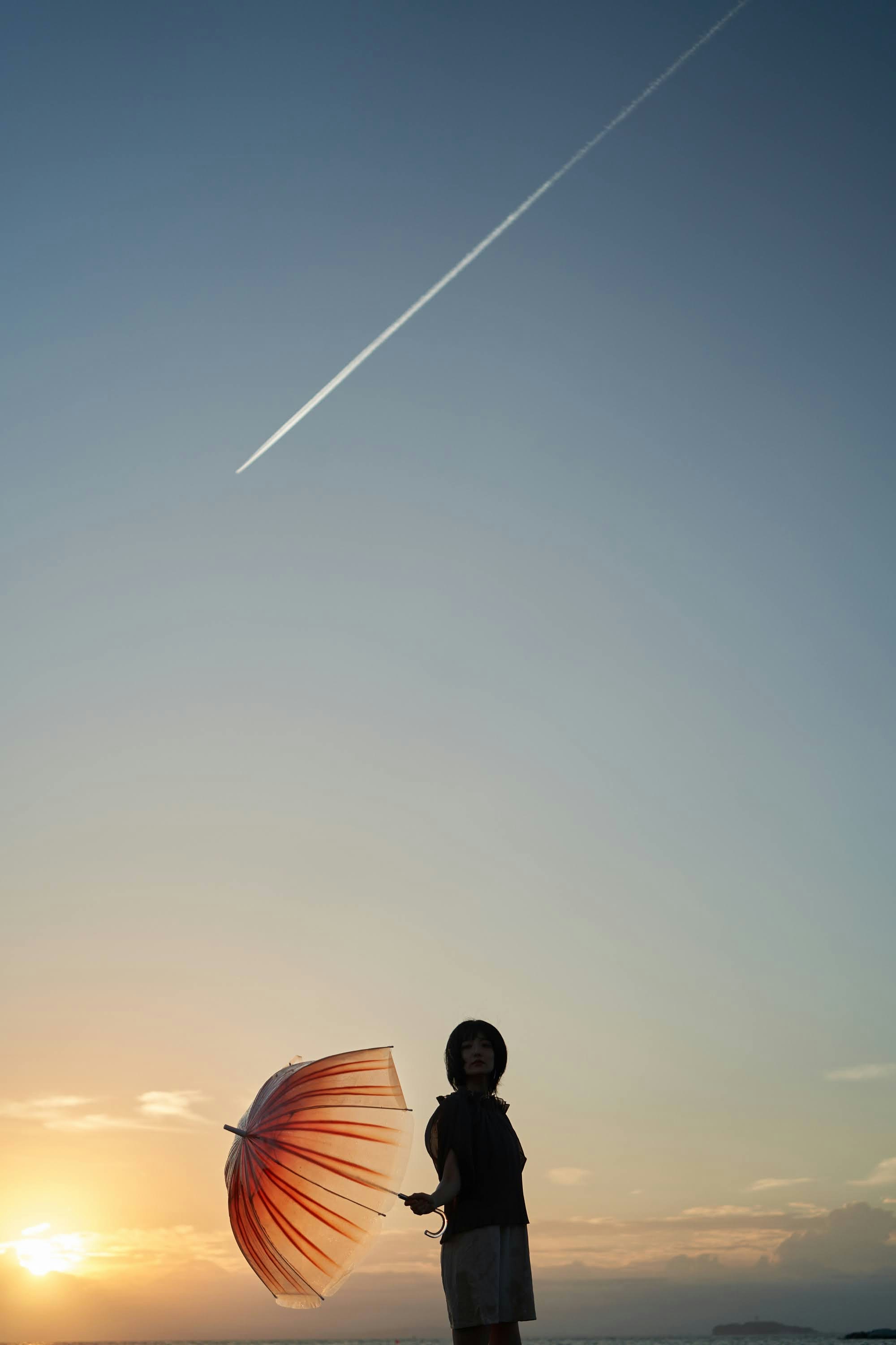 夕日を背景に飛行機雲と傘を持つ人物