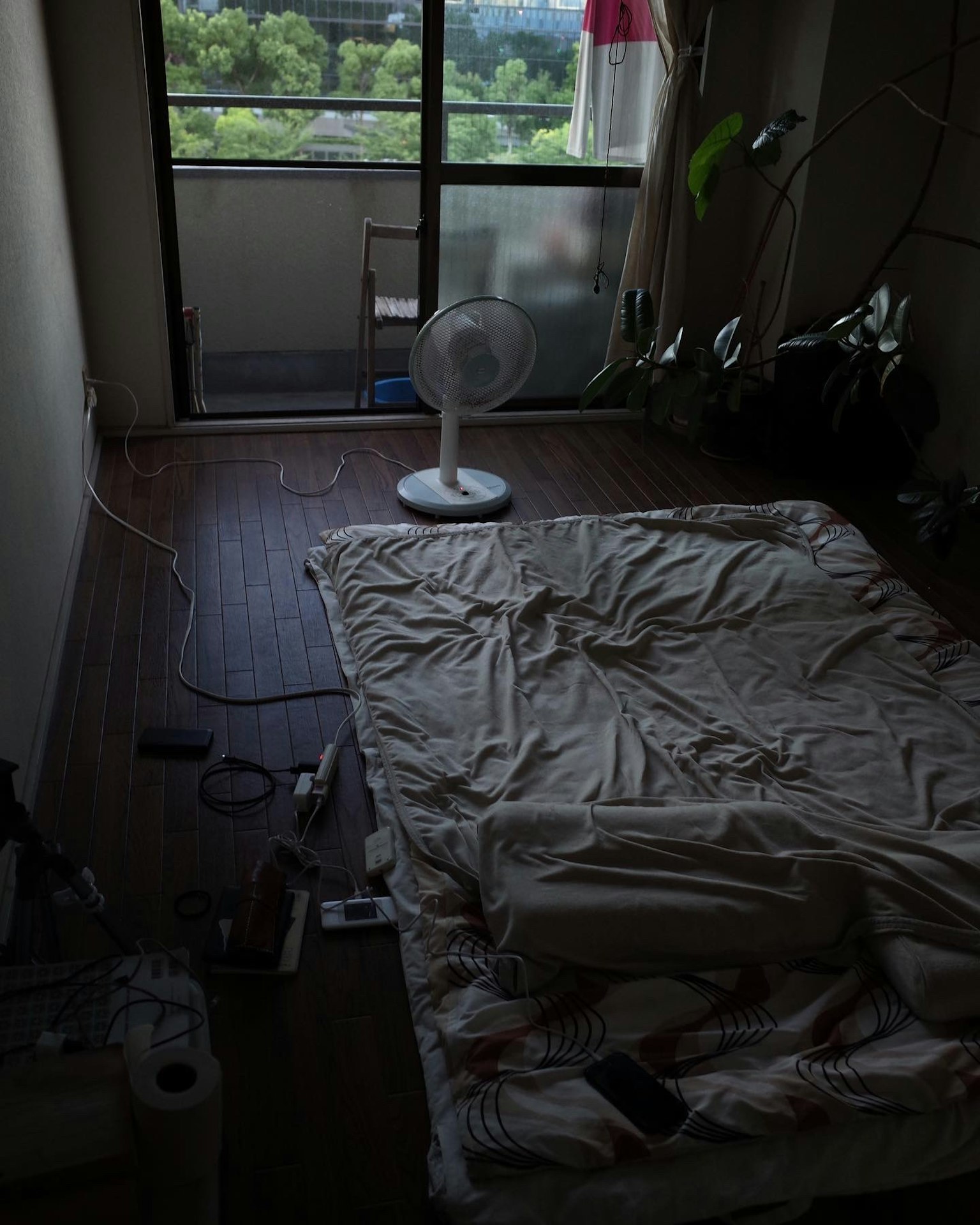 간단한 방 내부에 매트리스와 선풍기가 보이며 창문을 통해 녹지가 보인다