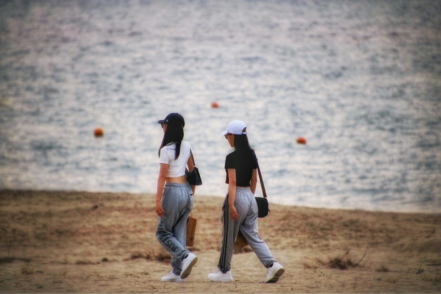 两位女性在海滩上走，一位穿着白色上衣，另一位穿着黑色上衣，搭配休闲裤，背景是大海