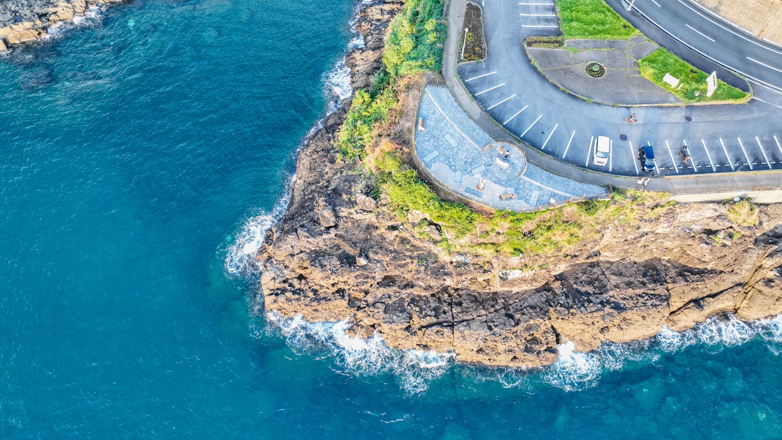 Vue aérienne d'une falaise rocheuse avec des vagues océaniques bleues à proximité comprenant un parking et des espaces verts