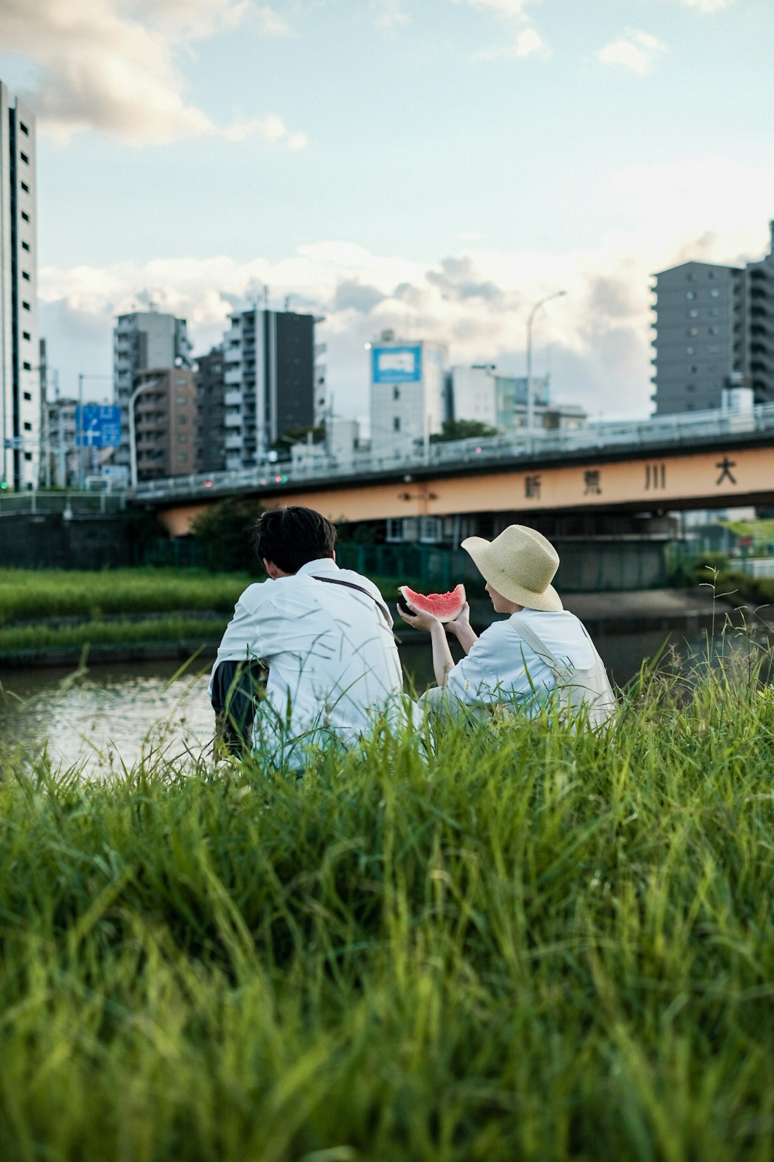Two people sitting by a riverside with skyscrapers in the background
