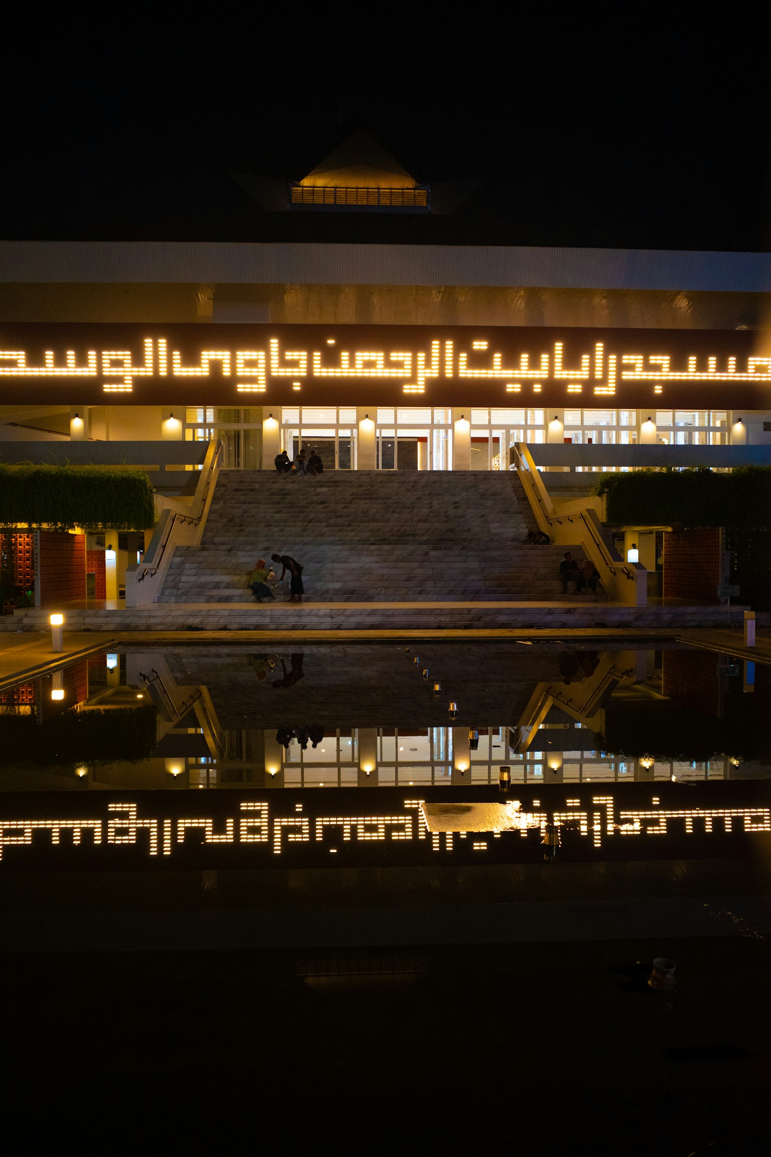Extérieur d'un bâtiment moderne illuminé la nuit se reflétant dans l'eau avec du texte arabe