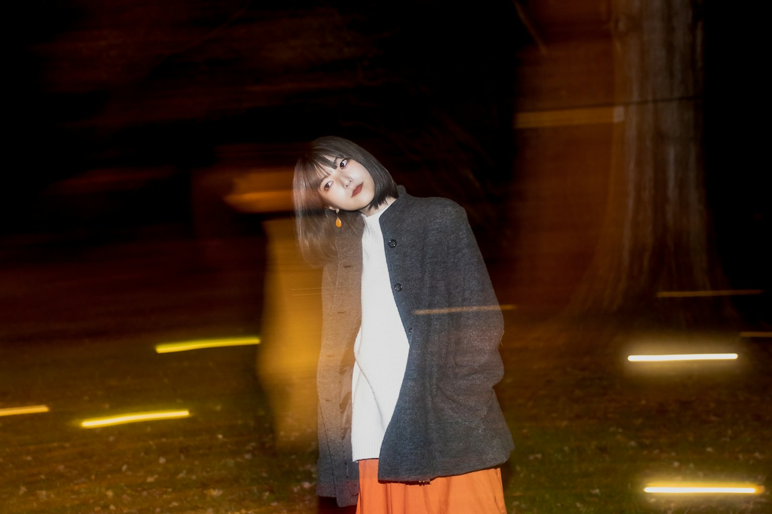 一位穿著橙色裙子的女性在夜晚的公園中站立，背景中有模糊的燈光