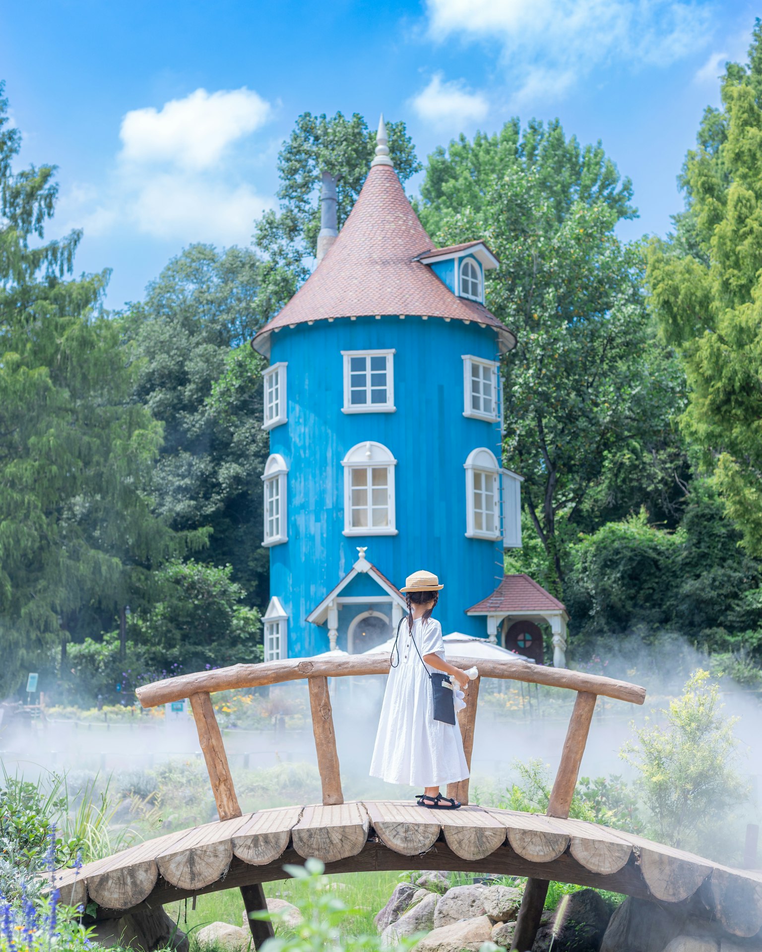 Une femme en robe blanche se tenant sur un pont avec une maison tour bleue en arrière-plan