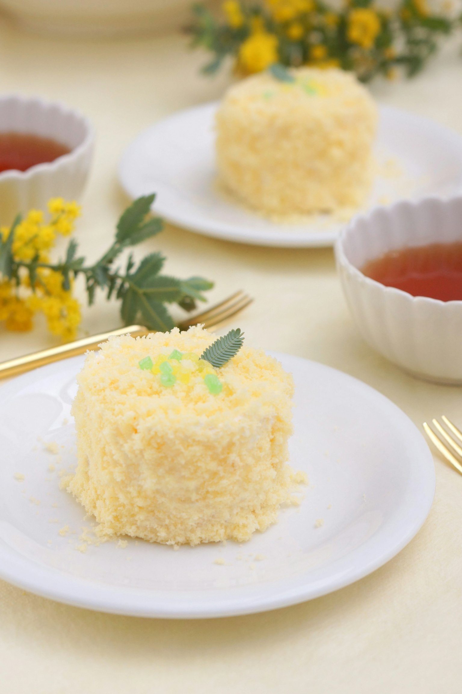 Gelber Kuchen auf einem weißen Teller mit Teetassen und Blumen im Hintergrund