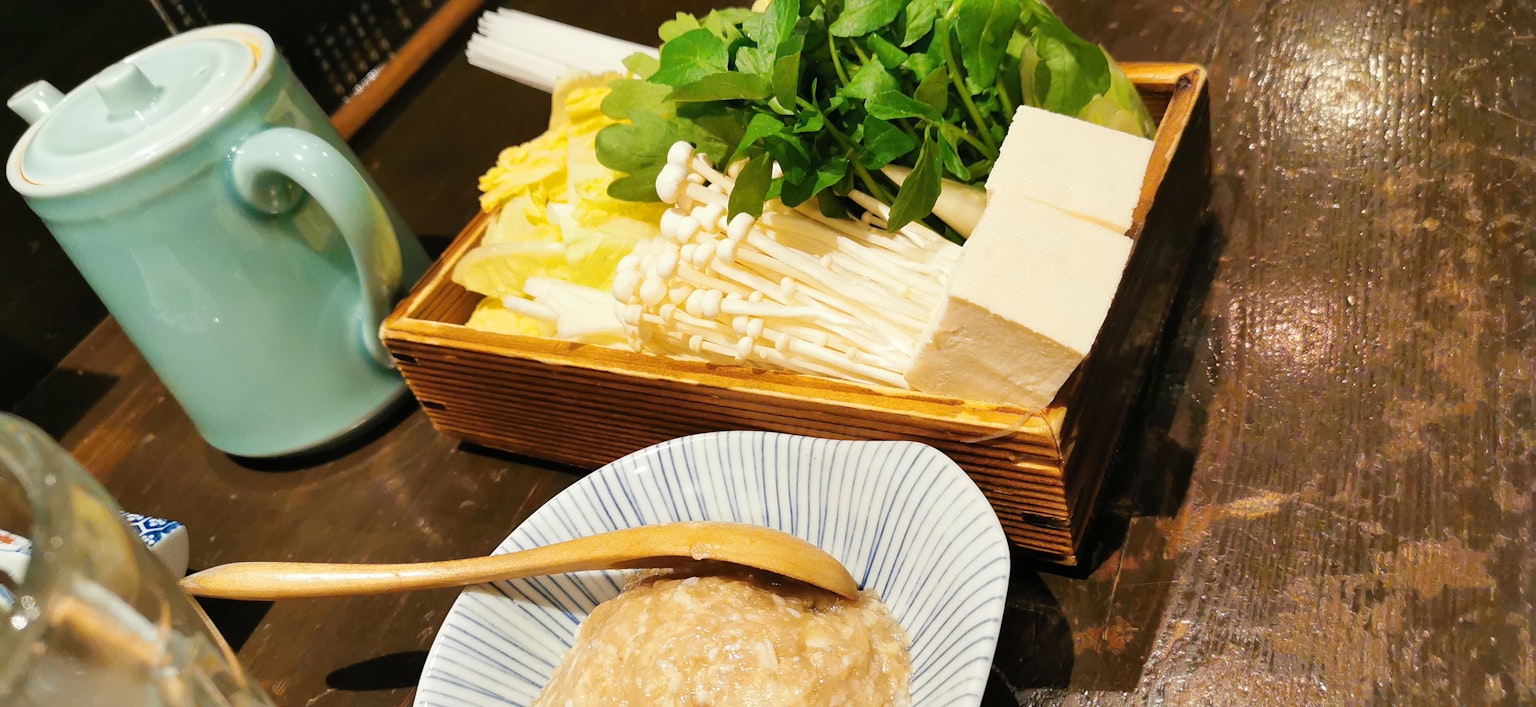 Một ấm trà màu xanh bên cạnh khay gỗ với rau củ và đậu phụ đa dạng