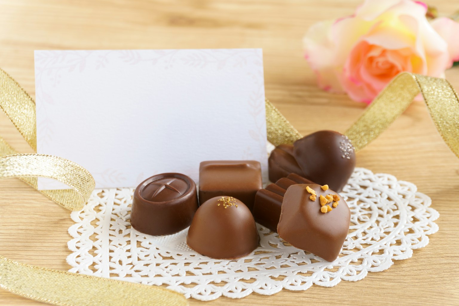 Assorted chocolate candies on a lace doily with a blank card and a pink rose
