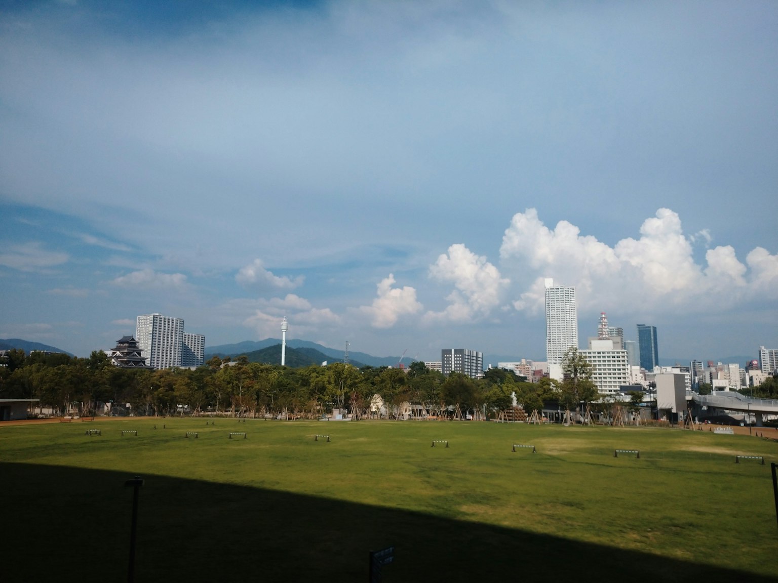 Pemandangan kota di bawah langit biru dengan awan putih menampilkan rumput hijau dan gedung pencakar langit