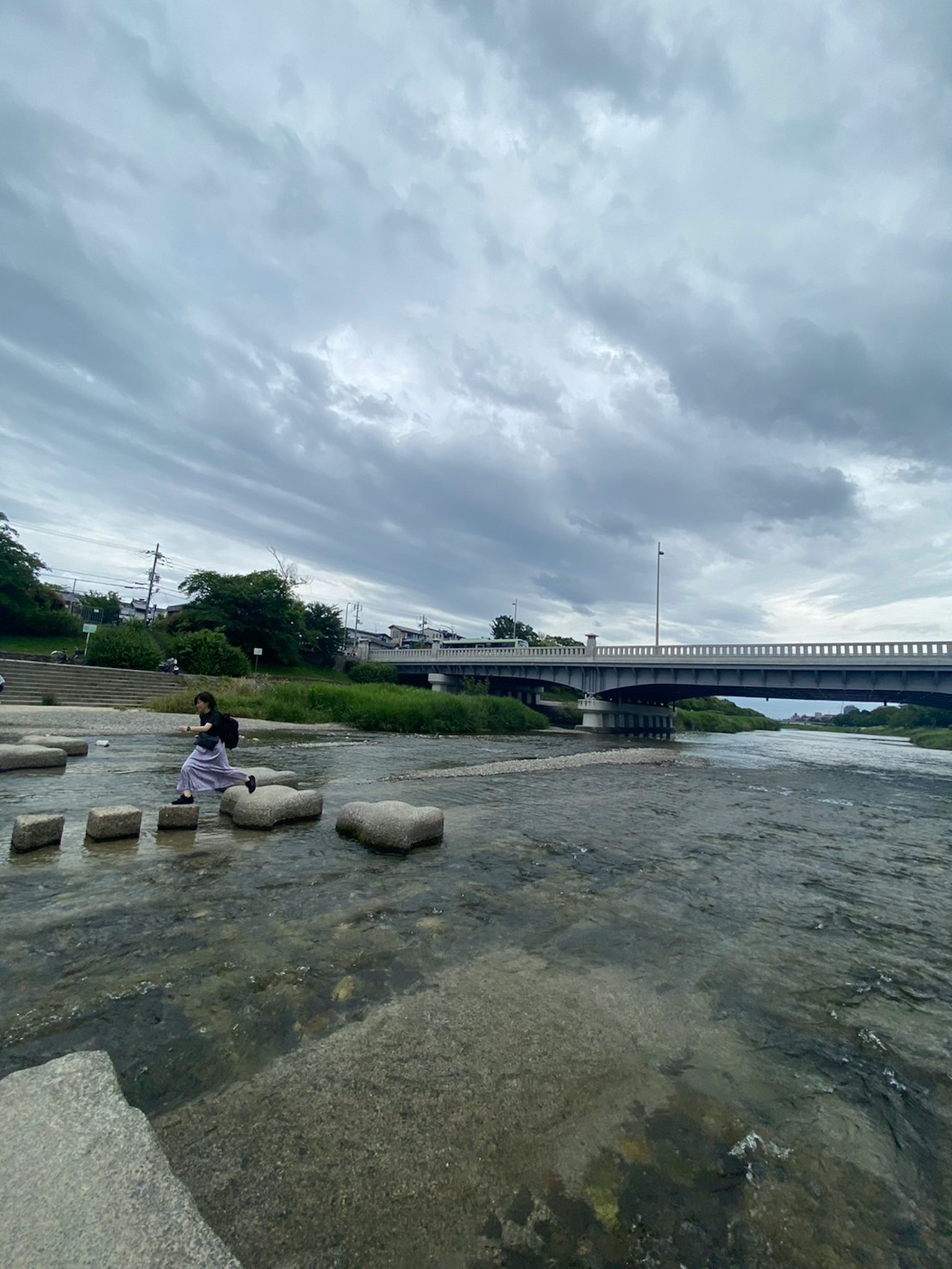 Enfant marchant sur des pierres dans une rivière avec un pont et un ciel nuageux