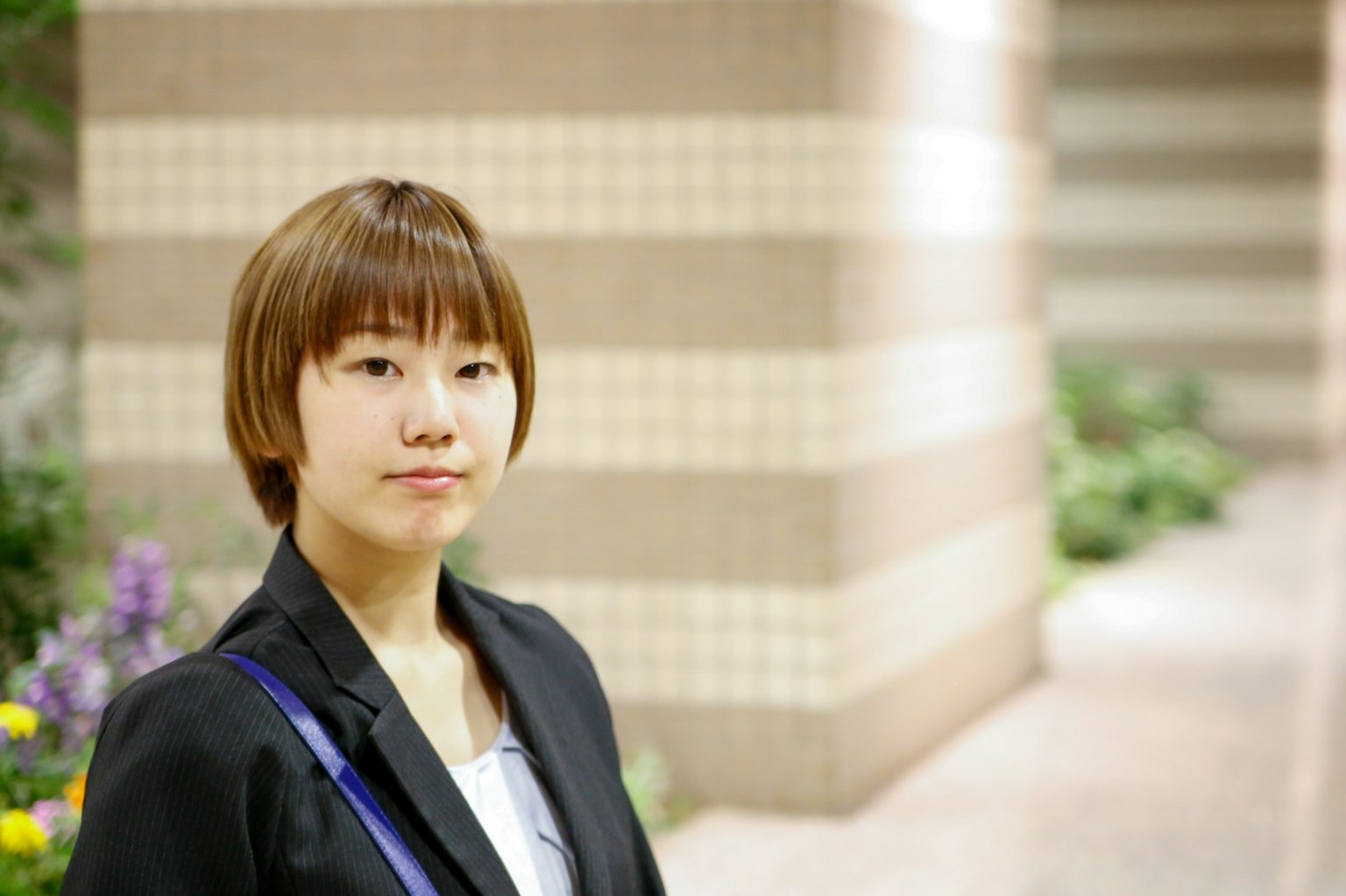 A woman in a business suit smiling with a tiled wall and plants in the background