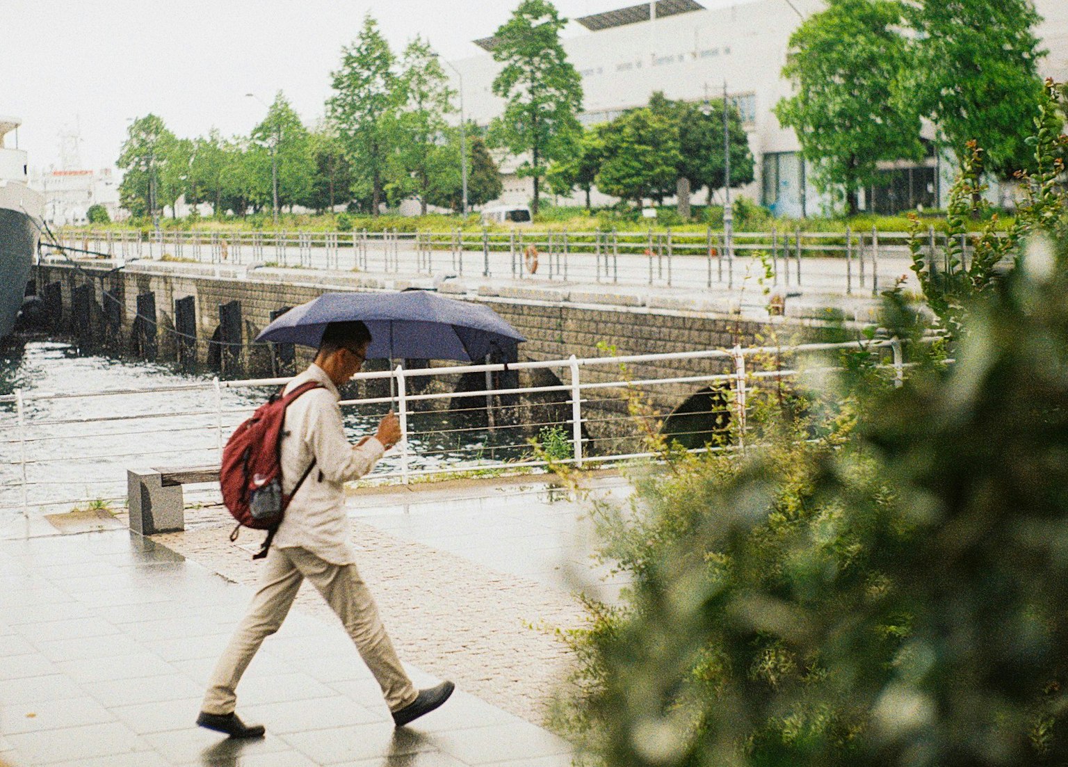 Ein Mann, der mit einem Regenschirm im Regen am Fluss entlanggeht, umgeben von grünen Bäumen