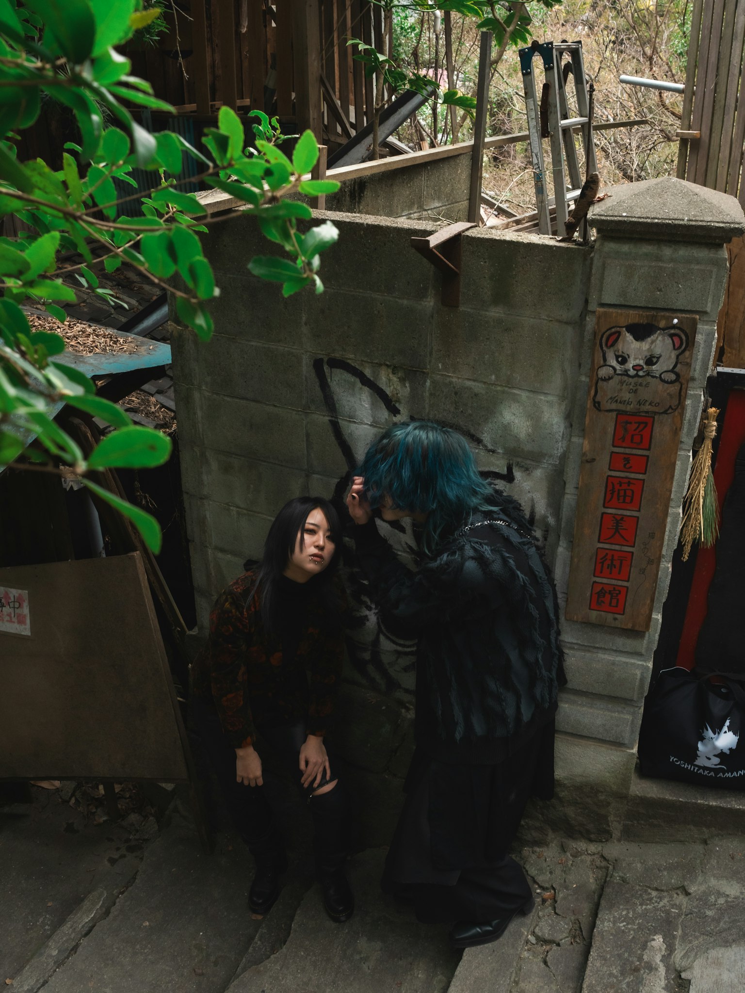 A woman sitting in front of an old wall surrounded by green leaves with a person adjusting her hair