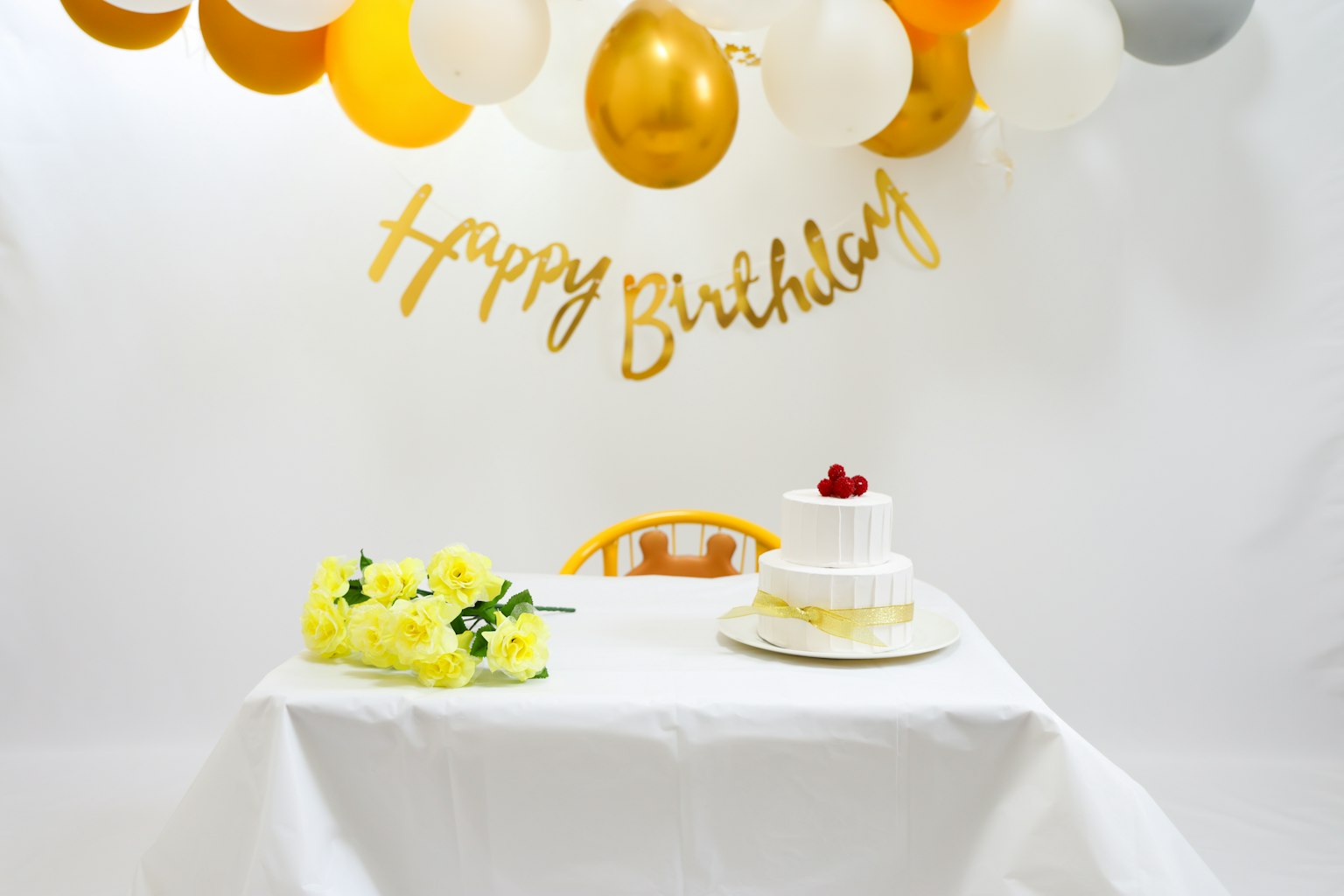 Une table blanche avec des fleurs jaunes et un gâteau à la crème avec une cerise sur le dessus, des ballons et une bannière Joyeux Anniversaire en arrière-plan