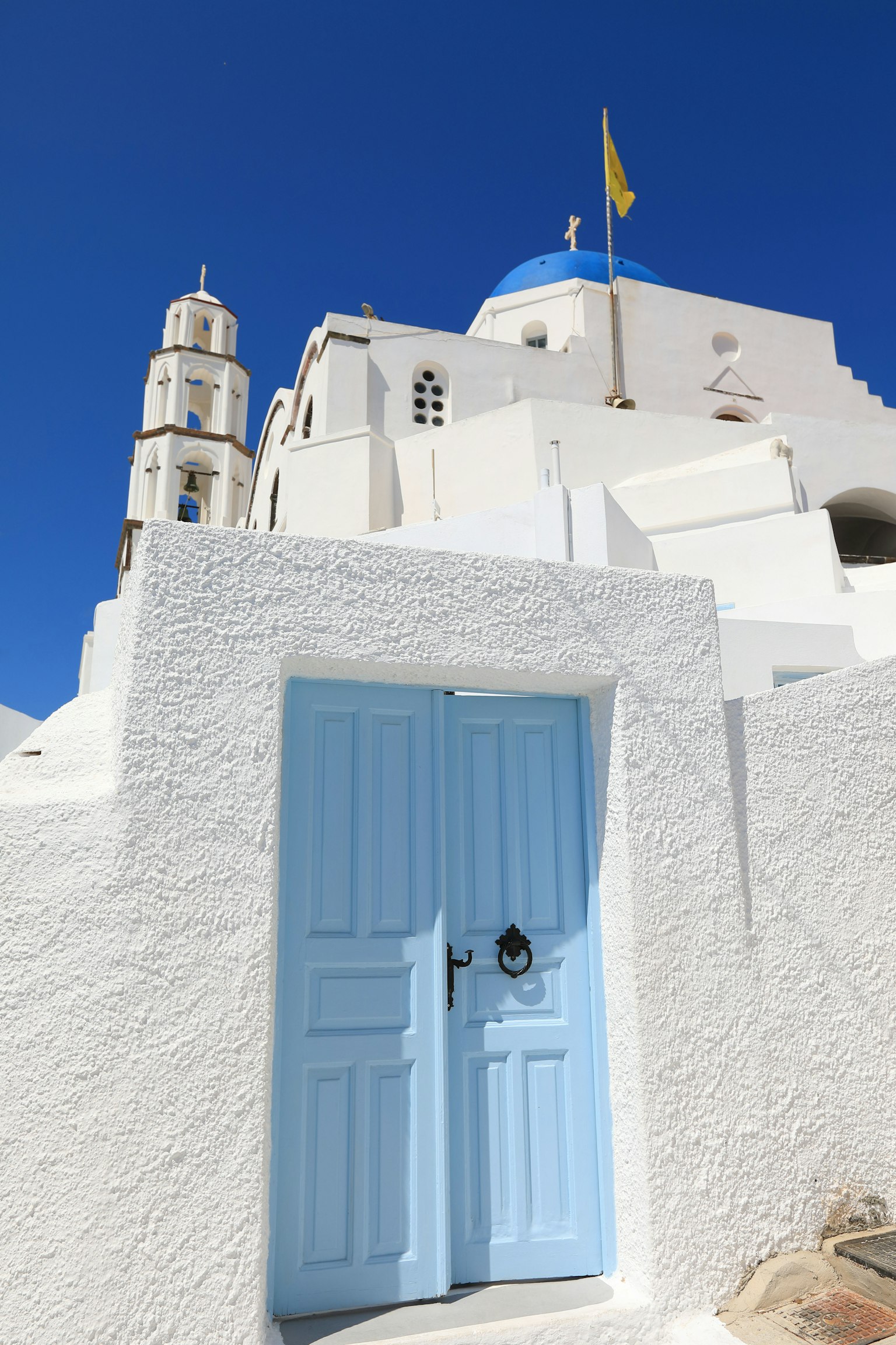 Khung cảnh đẹp với một tòa nhà trắng và cửa màu xanh, có tháp nhà thờ từ Santorini dưới bầu trời xanh rõ ràng