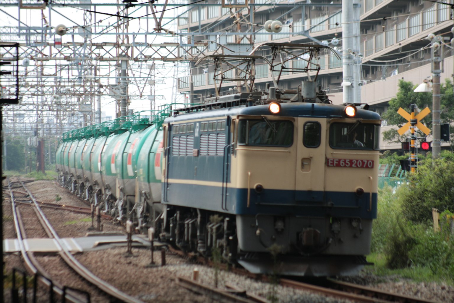 รถไฟสินค้าสีฟ้าและขาวลากรถถังสีเขียวในสภาพแวดล้อมในเมือง